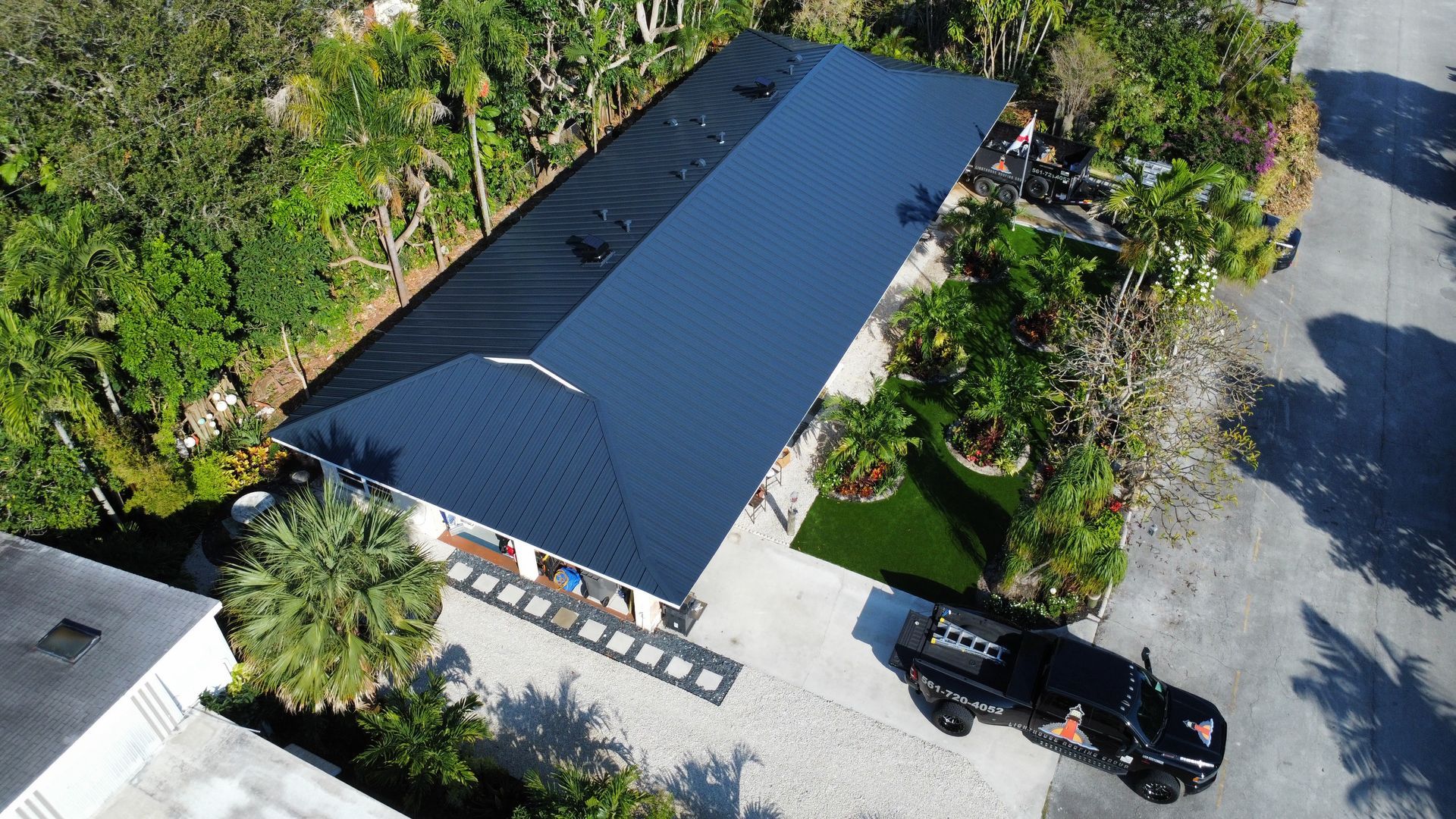 Aerial view of a long dark-roofed building with a paved driveway, parked car, and surrounding trees.