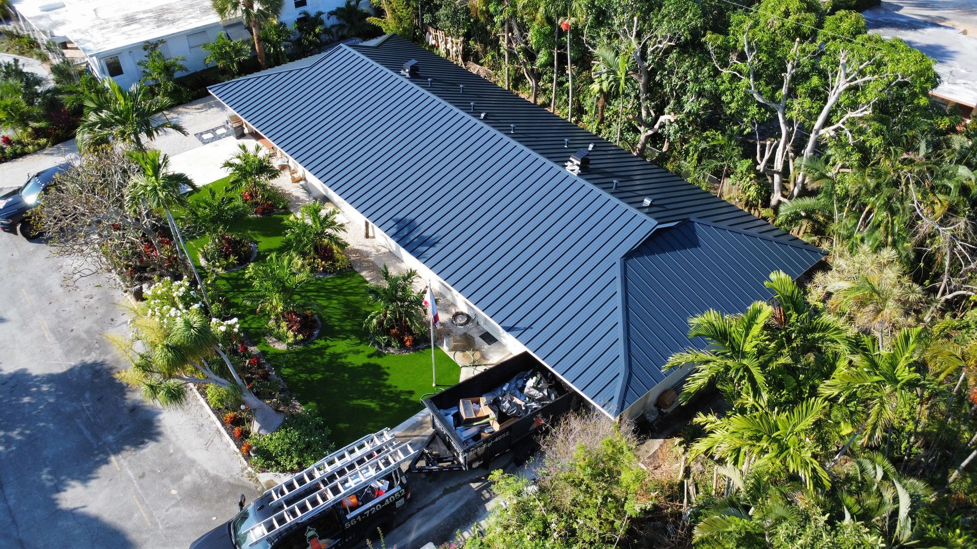 Aerial view of a modern house with a dark metal roof, surrounded by palm trees and greenery.