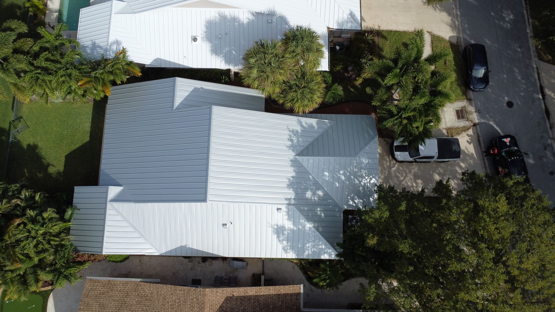 Aerial view of two white-roofed houses surrounded by trees, with a driveway and parked cars on the right.