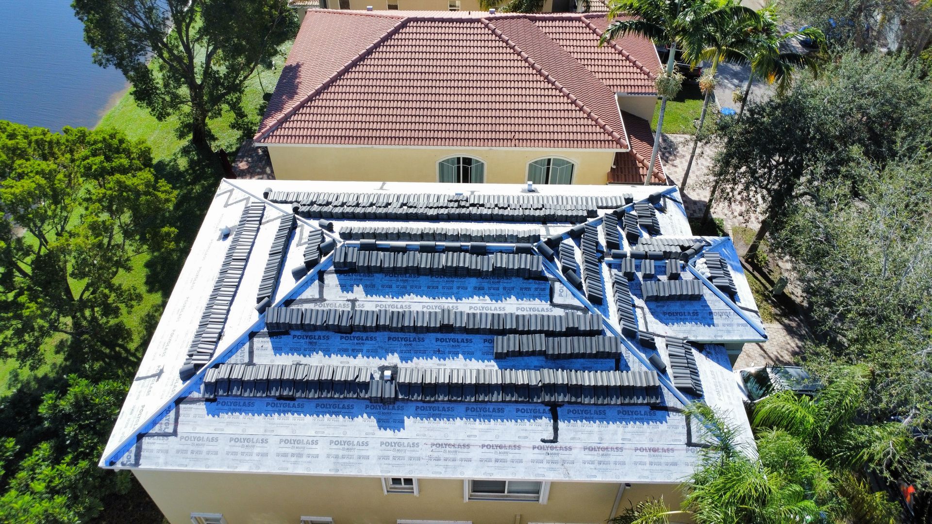 Aerial view of a white house with a red roof and a blue-tarp-covered roof area surrounded by trees