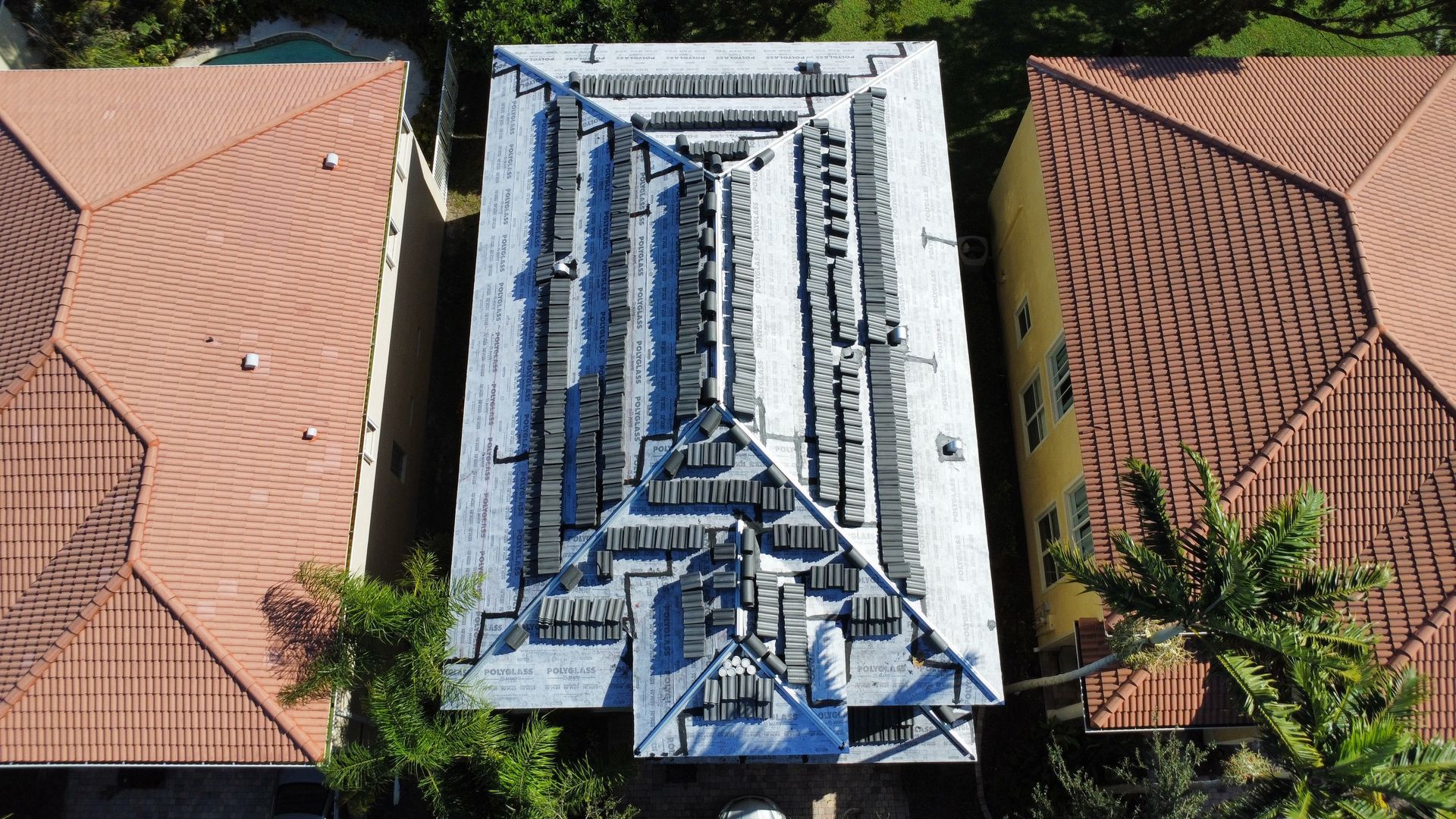 Aerial view of a white rooftop with blue solar panels between two red-tiled roofs and palm trees.