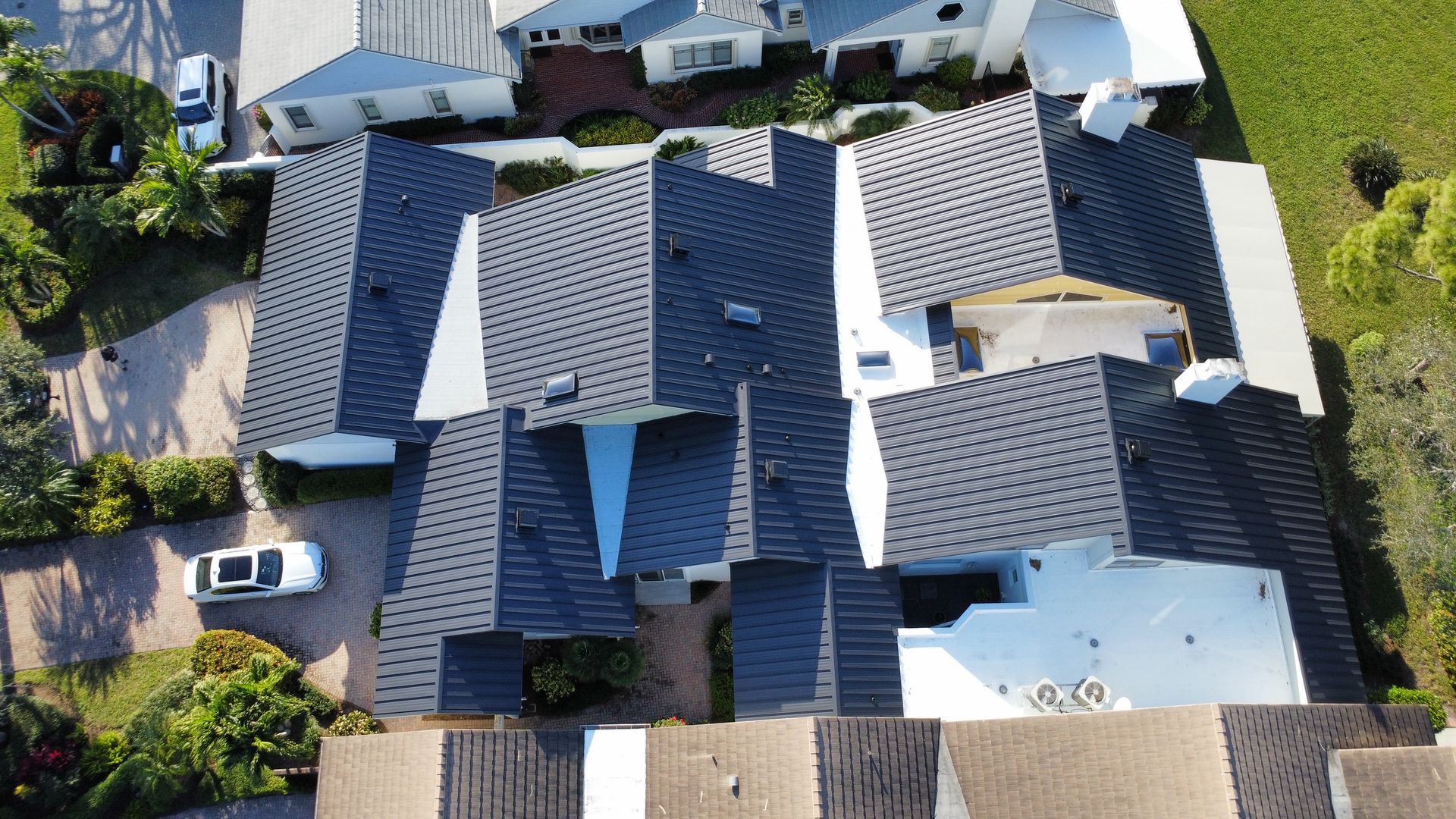 Aerial view of suburban houses with gray roofs, driveways, parked cars, and green lawns.