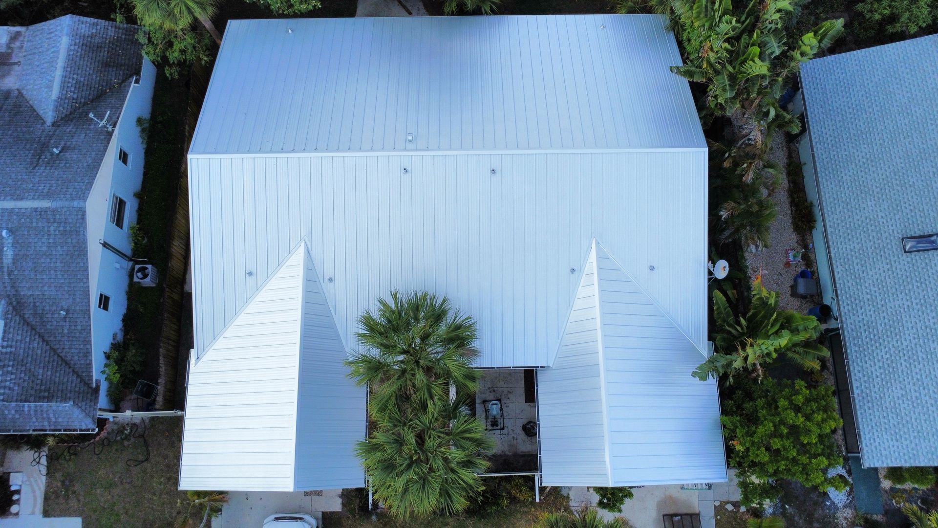 Aerial view of a white metal-roofed house with trees in the front yard, flanked by neighboring homes.