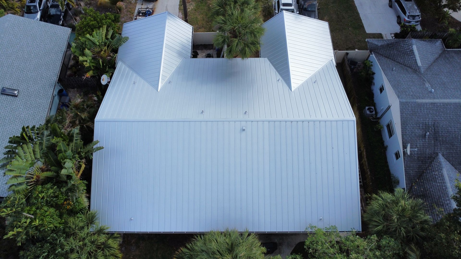 Aerial view of a large white-roofed house with two gables, surrounded by trees and neighboring homes.