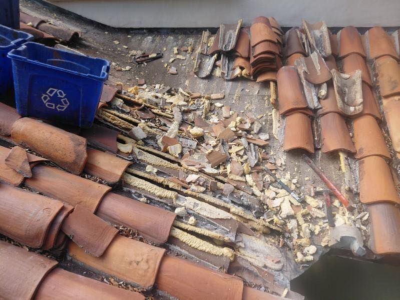 Damaged clay roof tiles with a blue recycling bin and debris scattered across the roof.