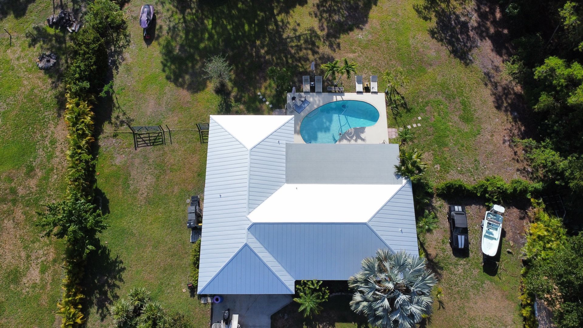 Aerial view of a white house with a blue-roofed pool, surrounded by trees and parked cars.