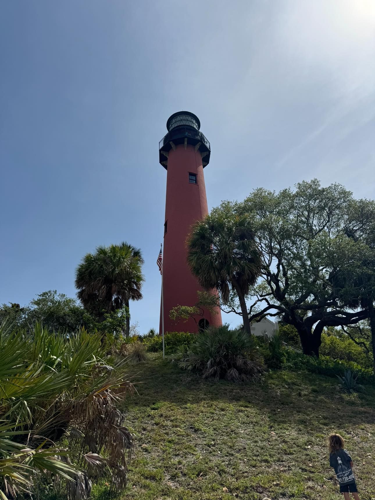 Tall red lighthouse with black top among green trees under a bright blue sky.