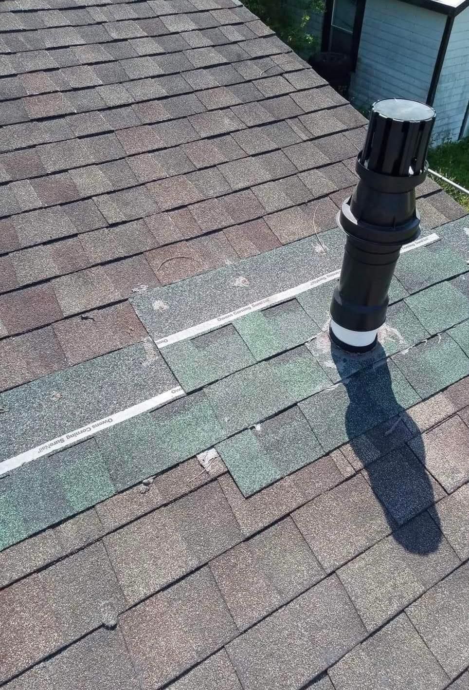 Rooftop vent pipe on weathered shingles with a patched section of green and brown roof tiles.