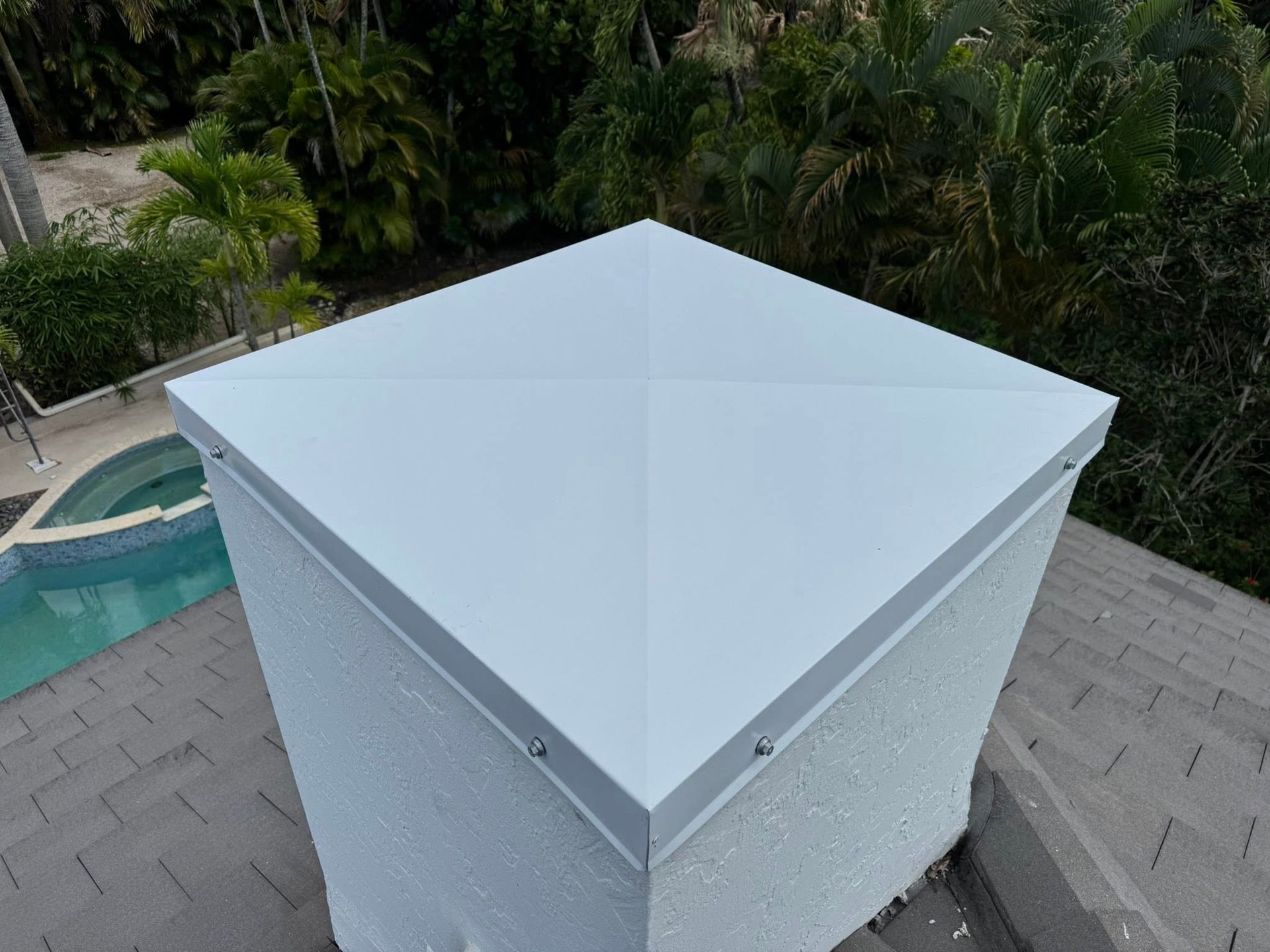 White rooftop vent on a gray shingled roof, viewed from above with trees in the background