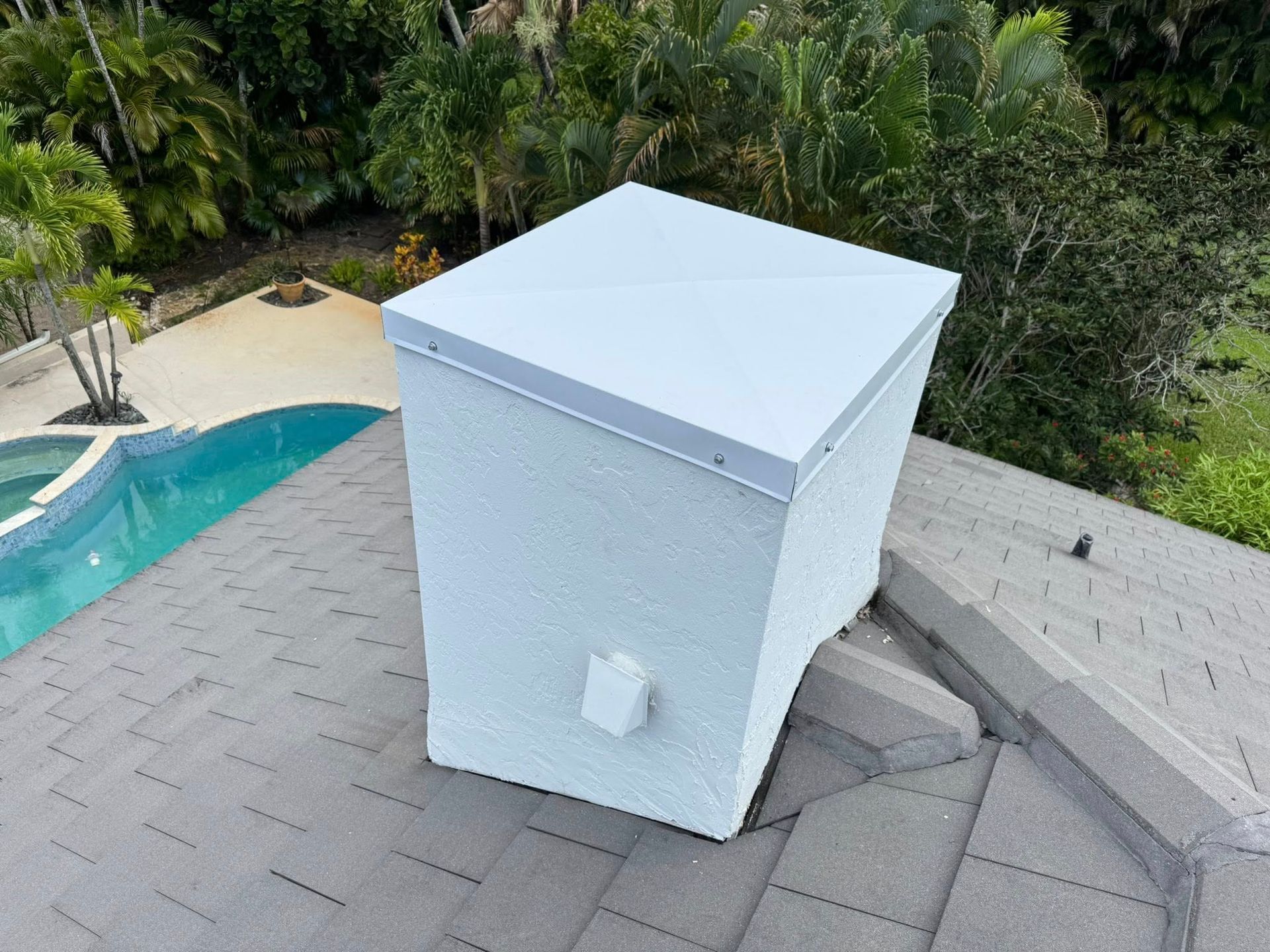 White roof vent on gray shingles beside a pool and tropical landscaping.