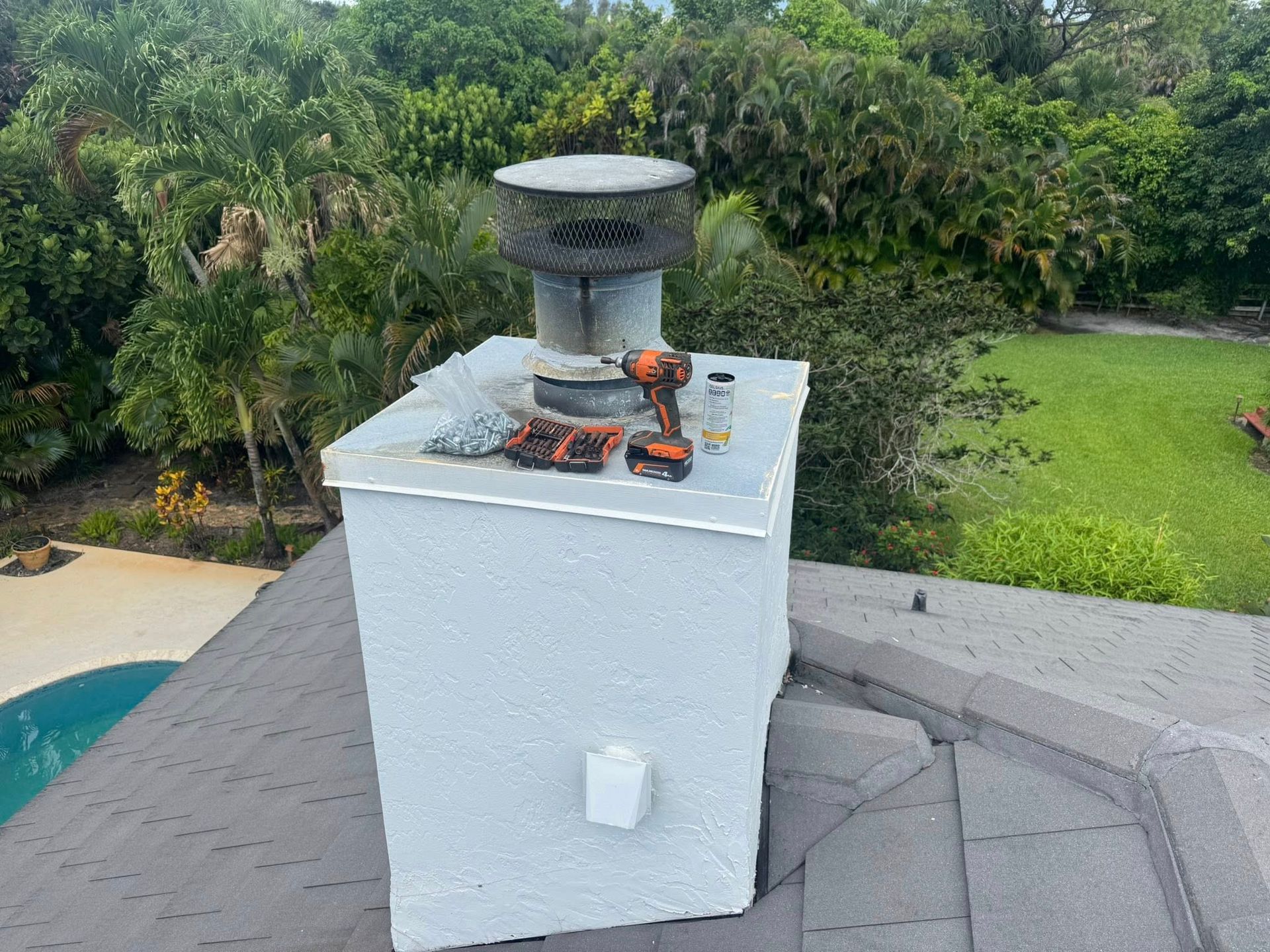 White rooftop chimney vent on a gray shingled roof with green yard and trees in the background