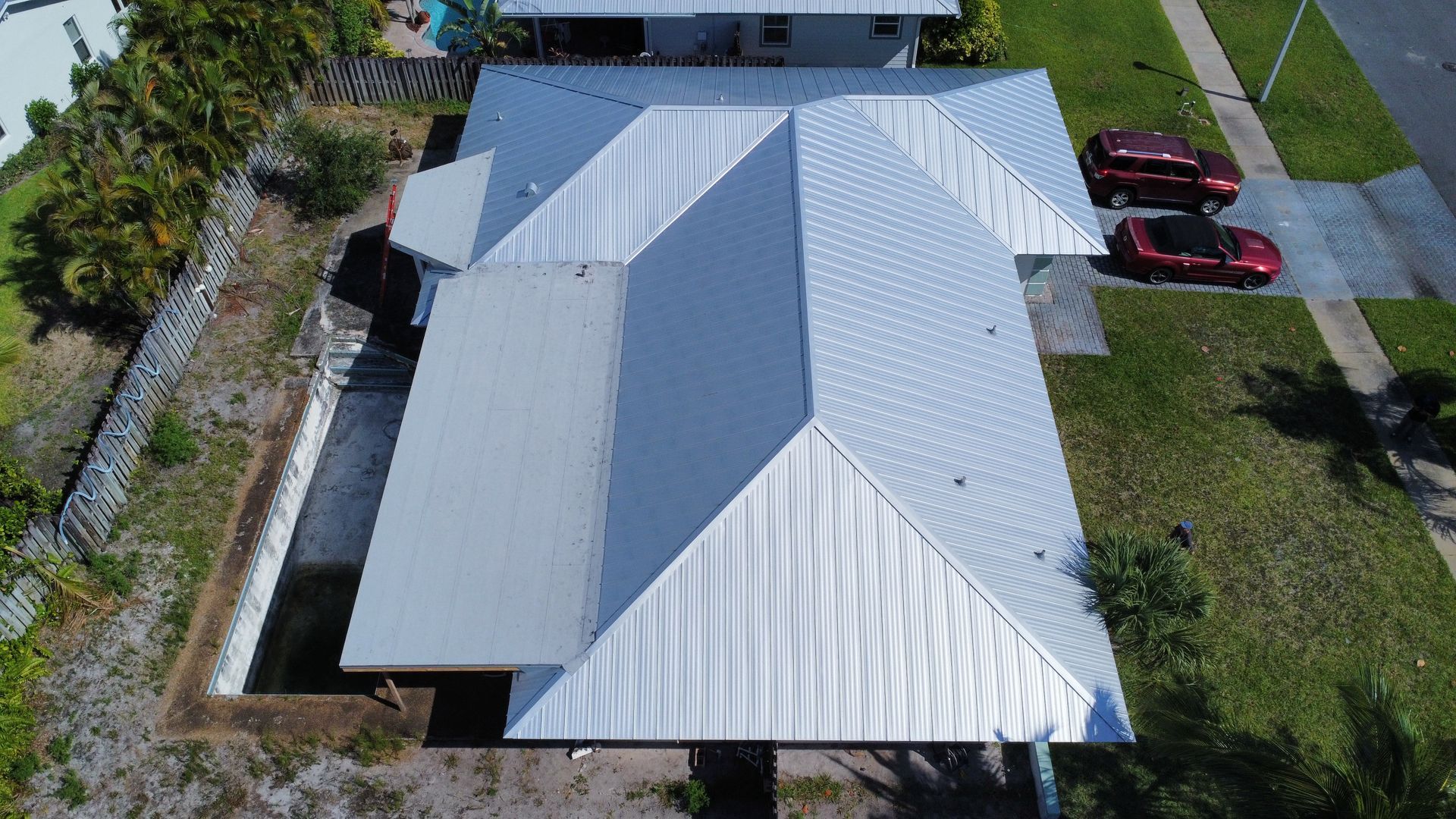 Aerial view of a house with a light metal roof, driveway, and two parked cars beside a grassy yard.