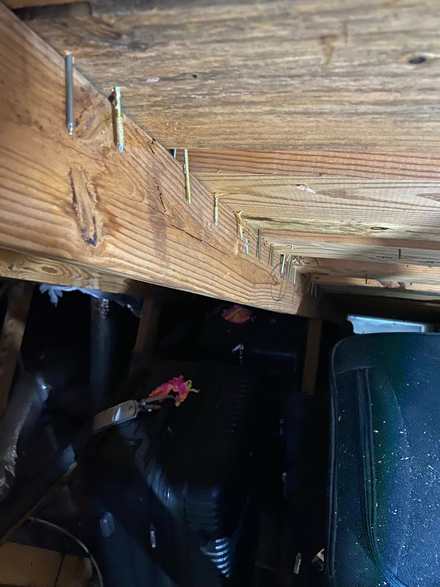 Close-up of a wooden beam with nails, above dark cluttered space and a black pipe or container below