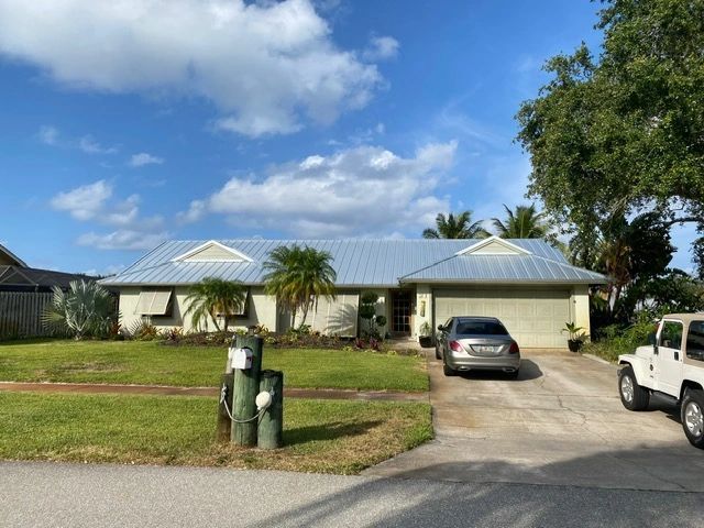 Single-story house with a white roof, driveway, parked cars, and a green lawn under a blue sky