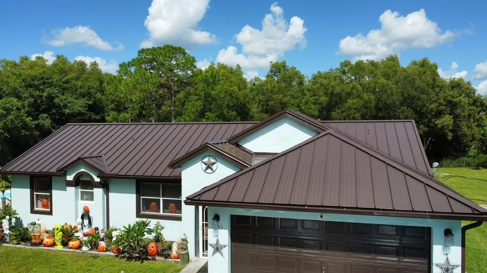 Single-story house with brown metal roof, white exterior, garage, and flower garden under a blue sky.