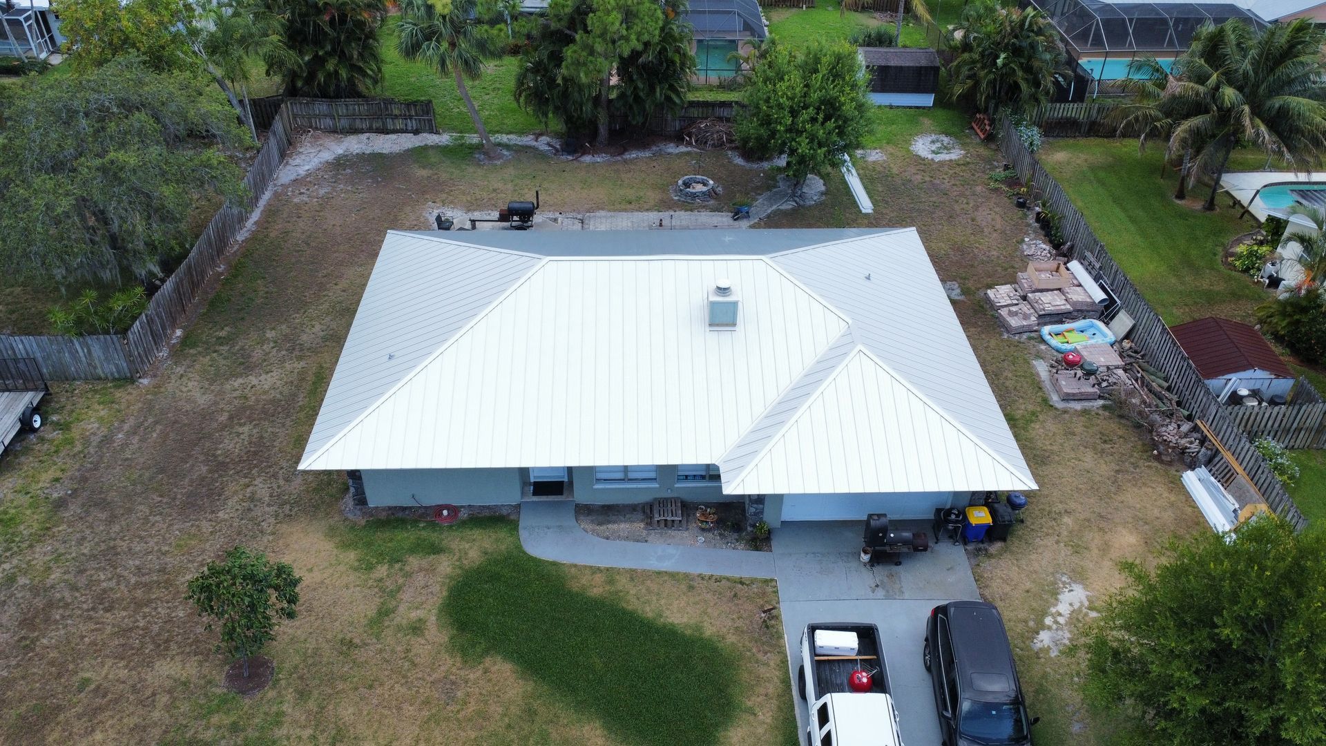 Aerial view of a white-roofed house with a driveway, yard, and surrounding trees