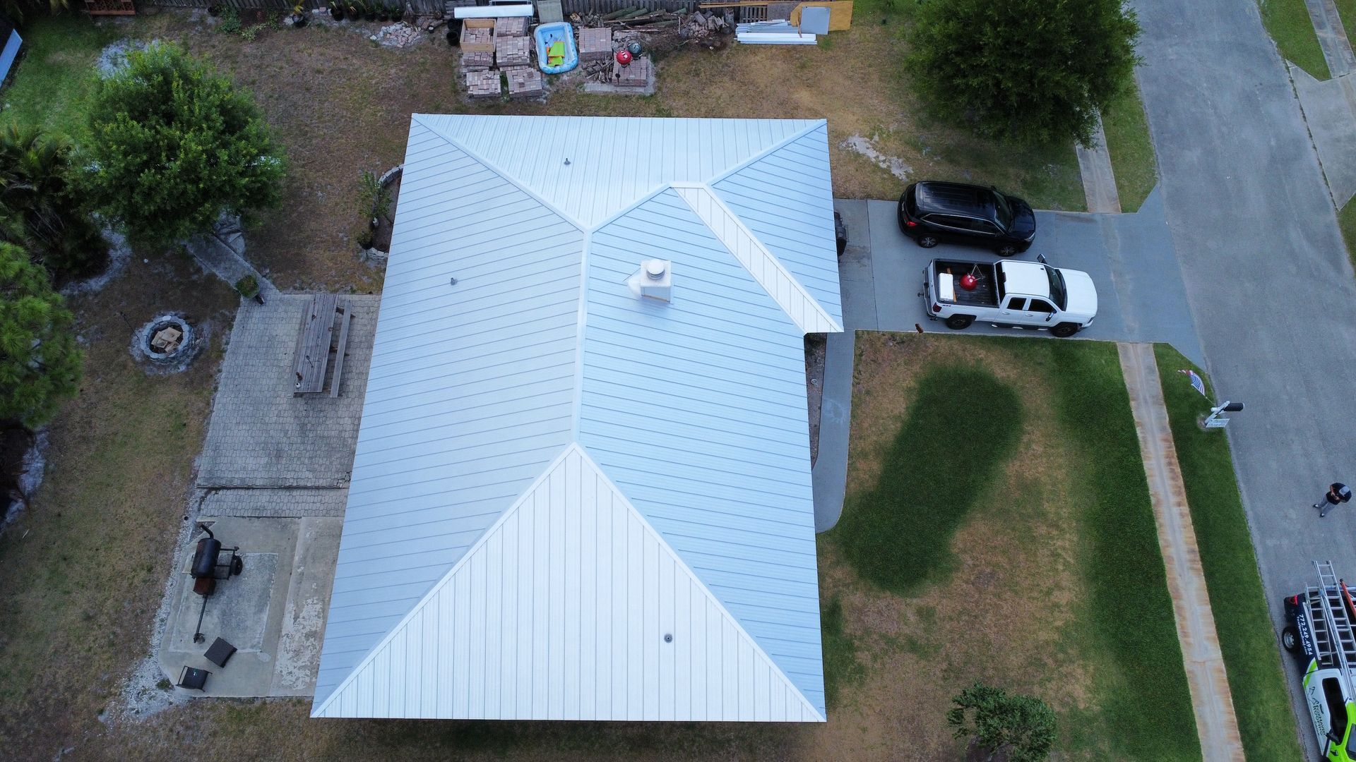 Aerial view of a white-roofed house with driveway, parked cars, and grassy yard.