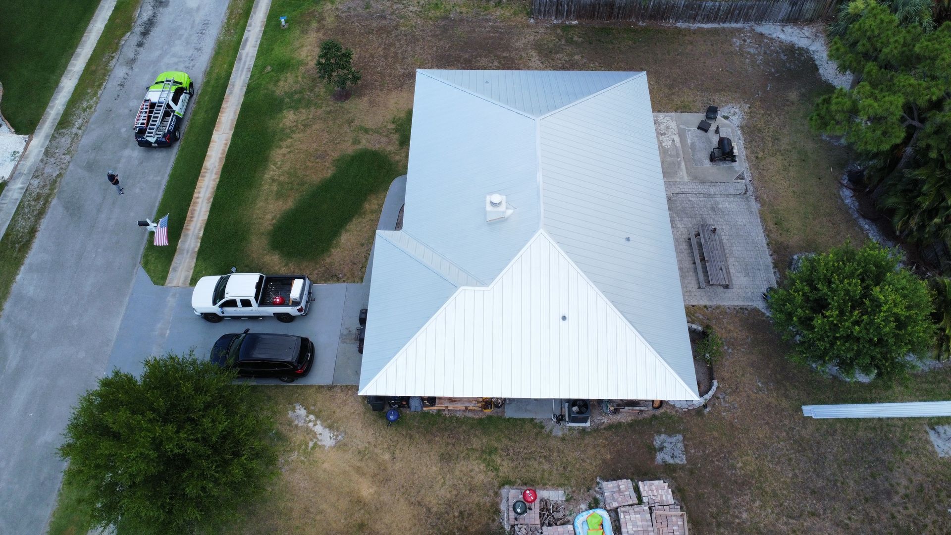 Aerial view of a white-roofed house with cars parked in the driveway and surrounding trees.