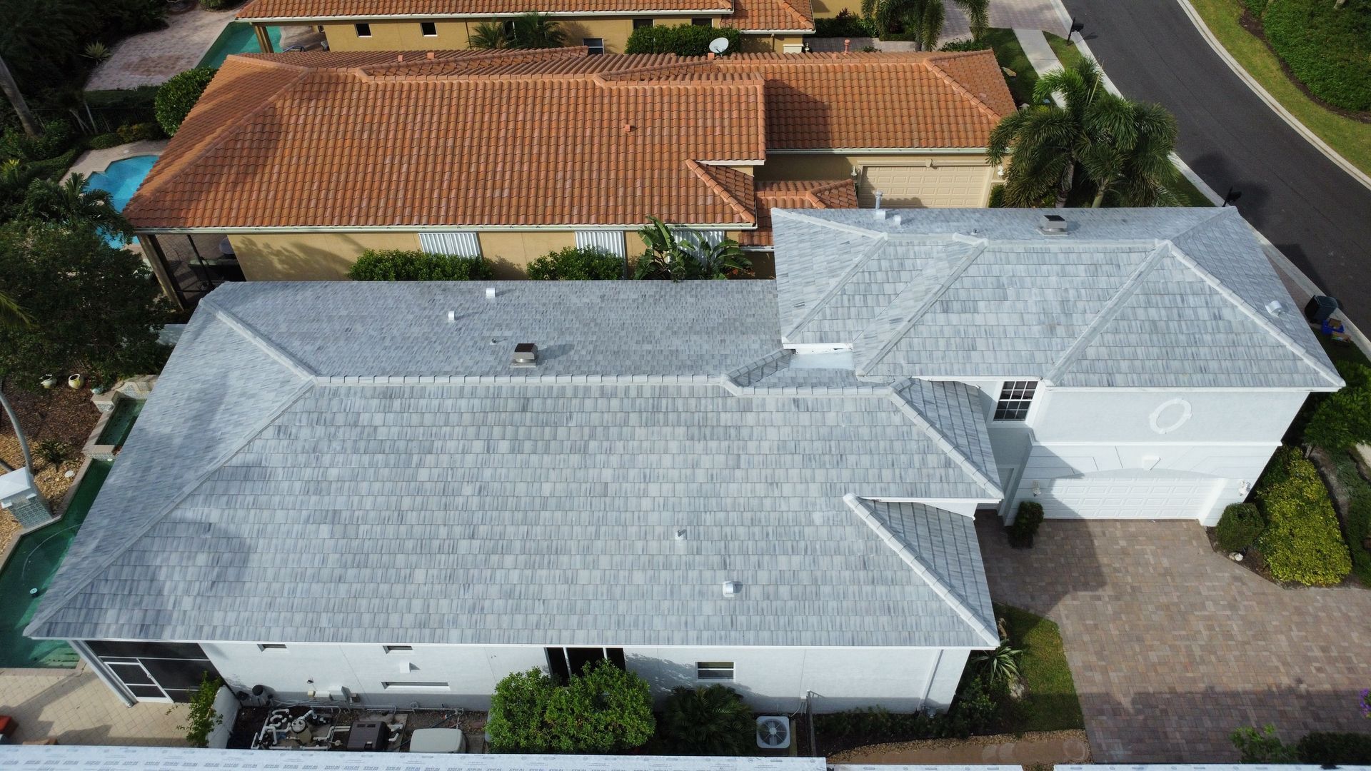 Aerial view of a light gray house with a driveway and adjacent orange-roofed homes in a suburban neighborhood