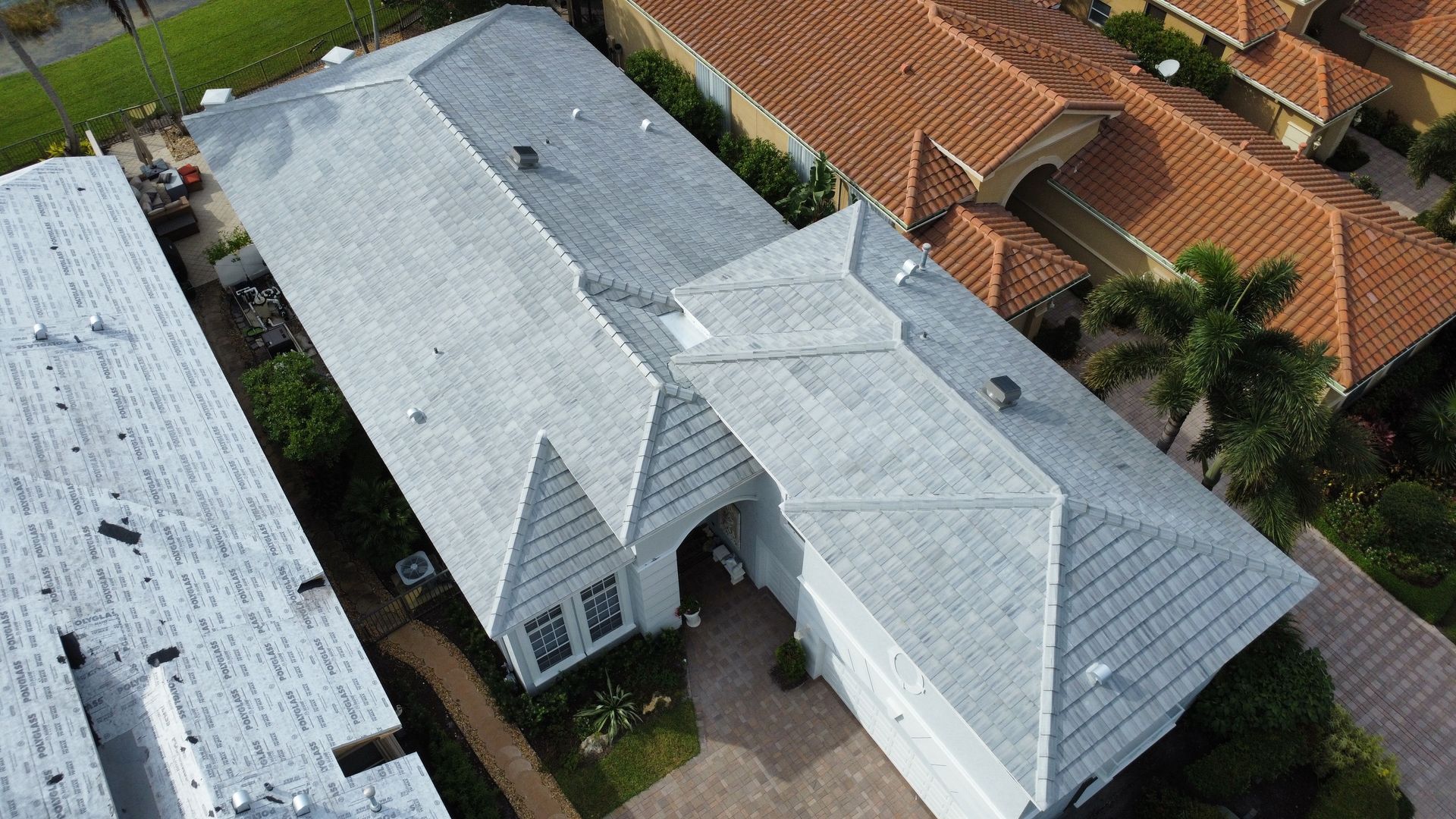 Aerial view of a white-roofed house with a driveway, beside orange-tiled neighboring homes.