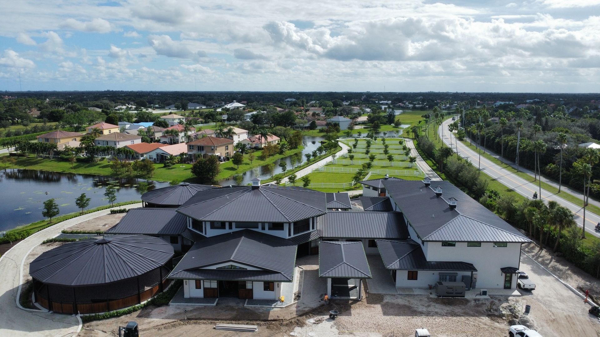 Aerial view of a modern black-roofed building complex beside a road, with trees, homes, and water nearby.
