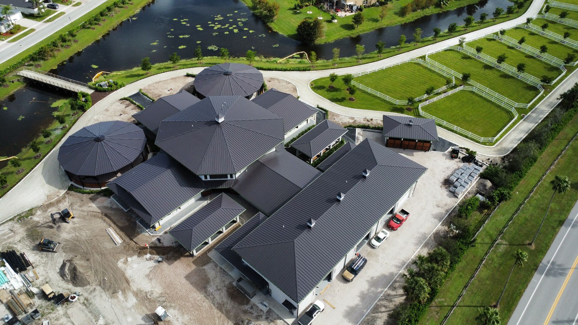 Aerial view of a large dark-roofed building beside a pond and road, with surrounding trees and lawns