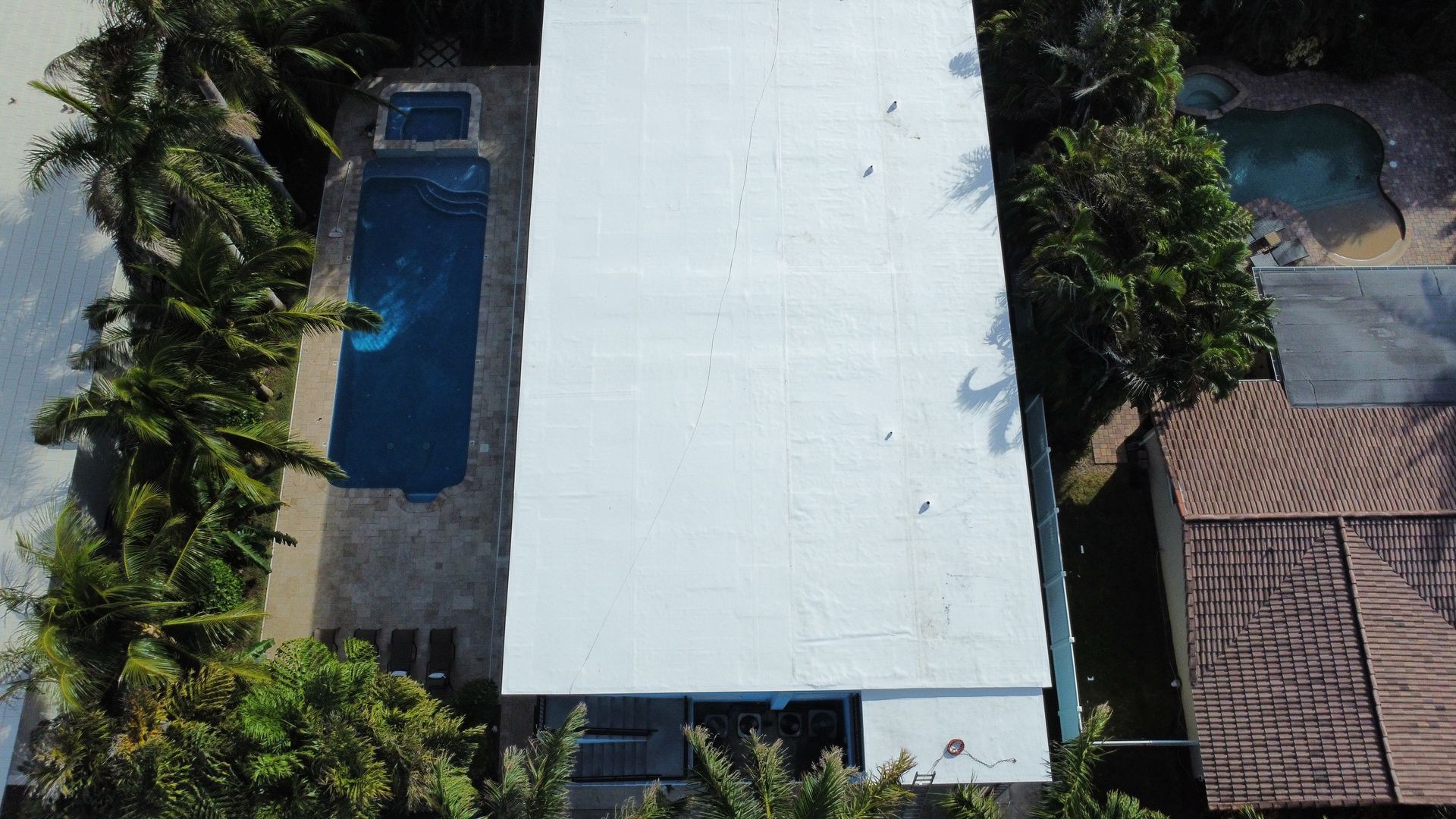 Aerial view of a white-roofed building beside a narrow blue swimming pool and palm trees.