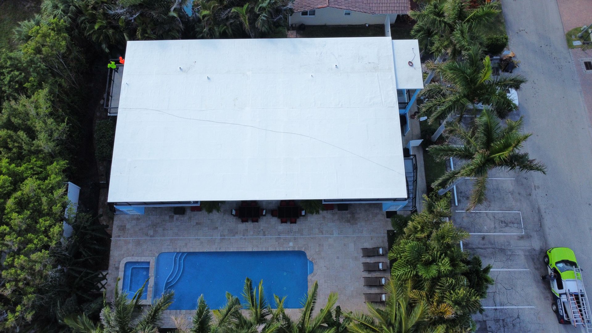 Aerial view of a house with a white roof, blue swimming pool, and palm trees beside a road.