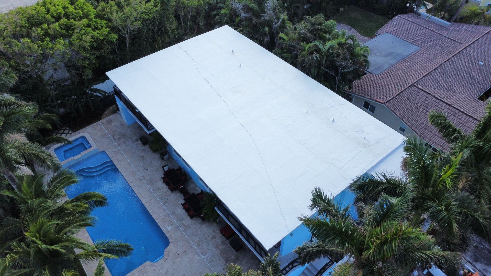 Aerial view of a large white-roofed house beside a blue swimming pool and palm trees.