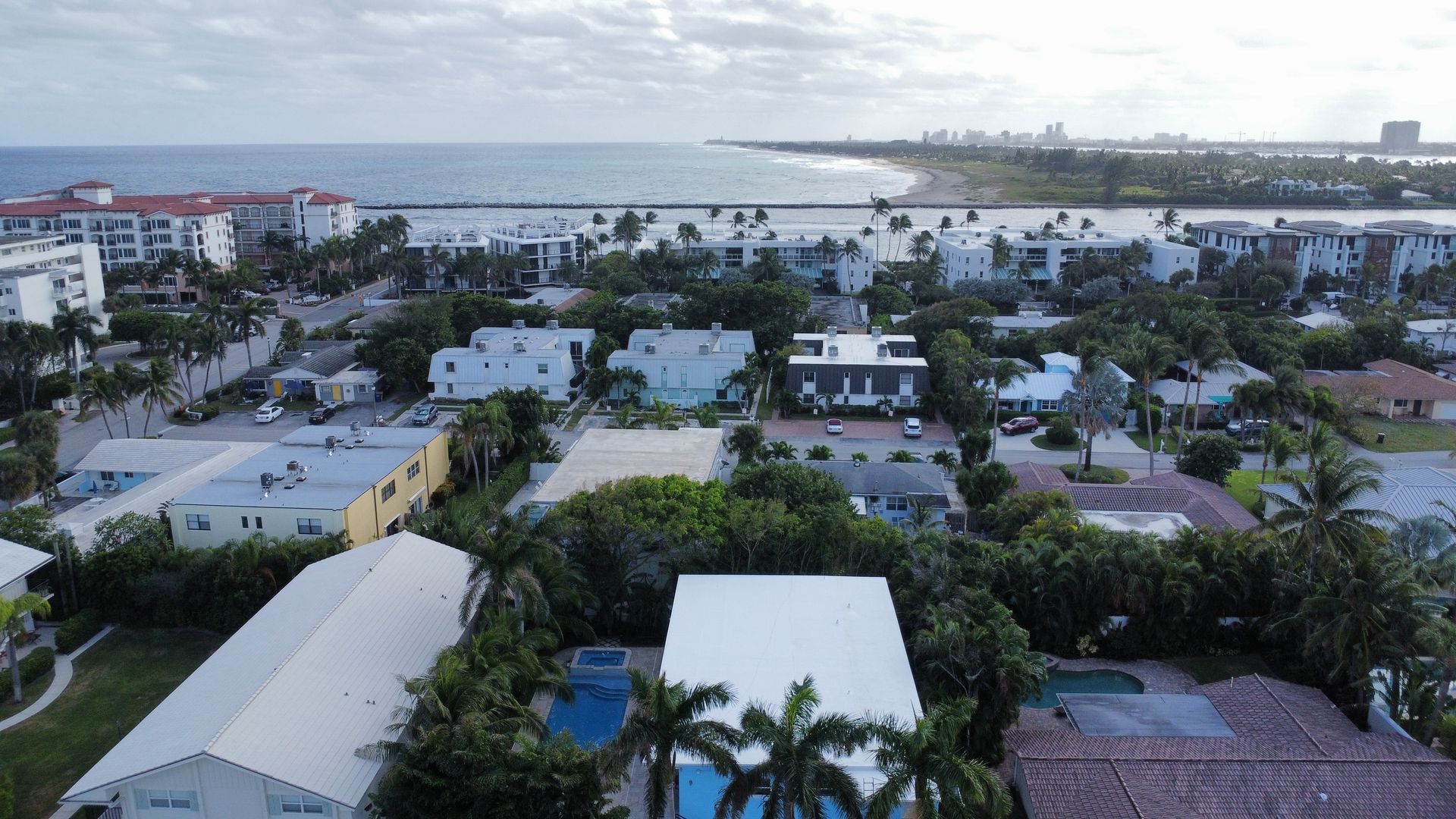Coastal neighborhood with white-roofed homes, palm trees, and water in the distance under a cloudy sky