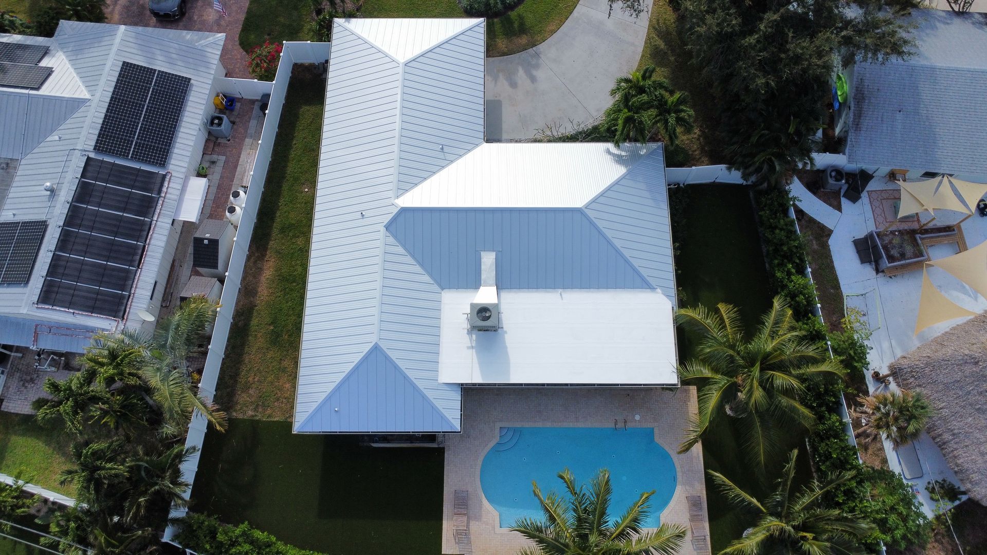 Aerial view of a white-roofed house with a blue pool, surrounded by palm trees and neighboring buildings