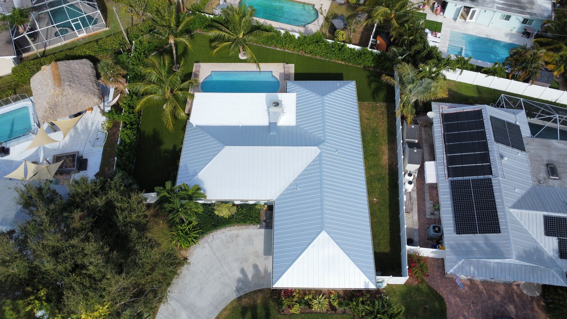 Aerial view of a white-roofed house with a driveway, surrounded by trees and neighboring homes.