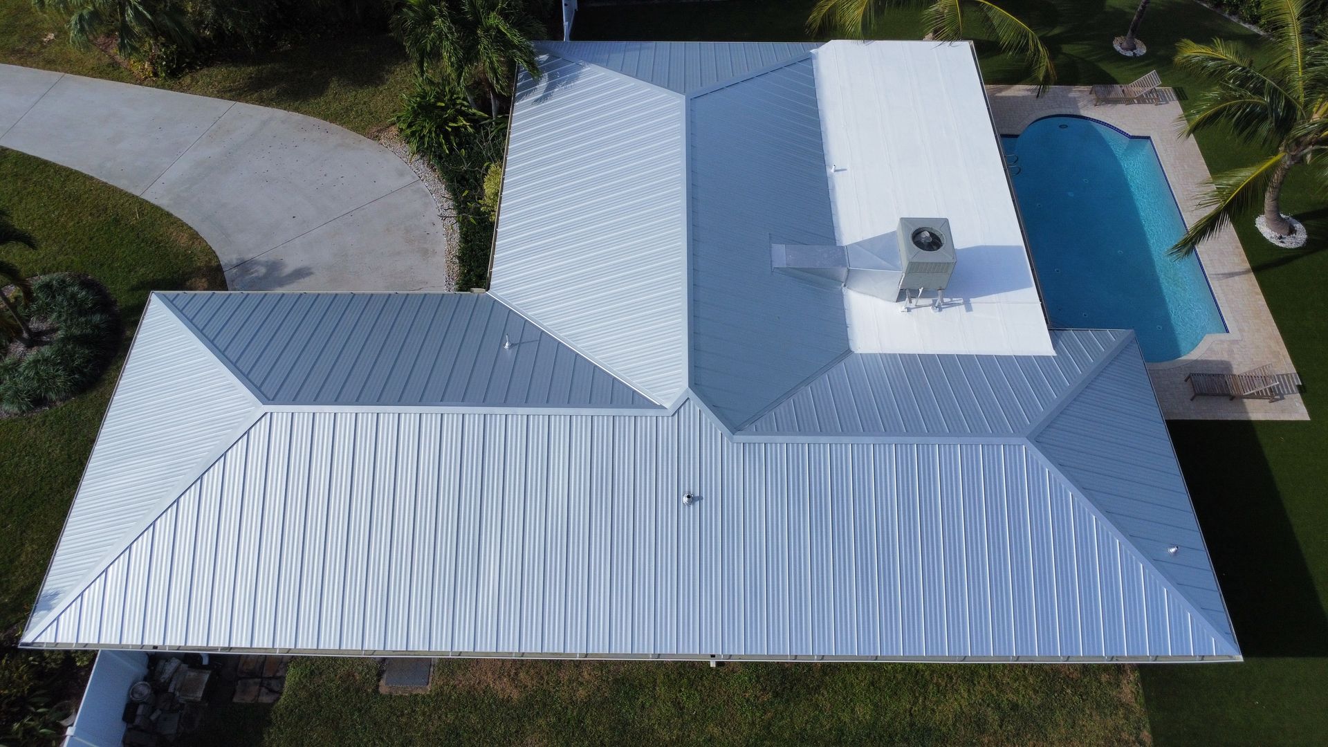 Aerial view of a white-roofed house with a driveway, lawn, and backyard pool.