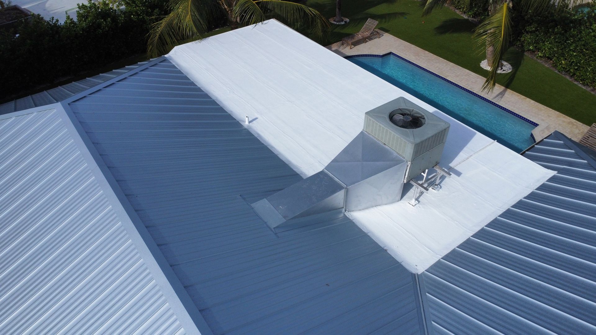 Aerial view of a white and blue metal roof with a central vent and duct near a poolside yard