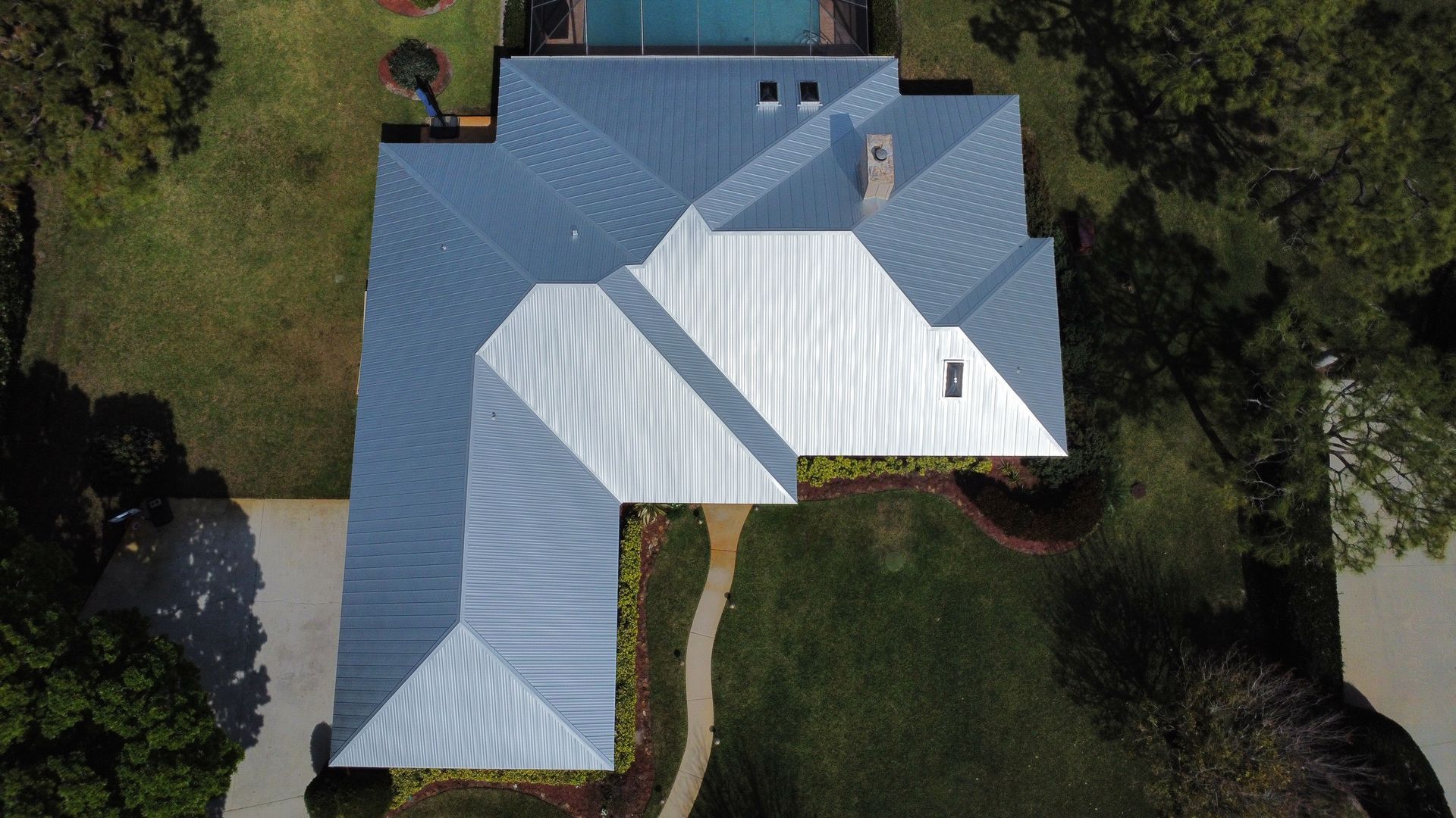 Aerial view of a house with a gray metal roof, surrounded by trees and a driveway.