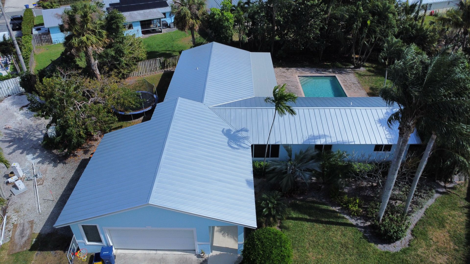 Aerial view of a white-roofed house with a covered patio and swimming pool, surrounded by tropical trees.