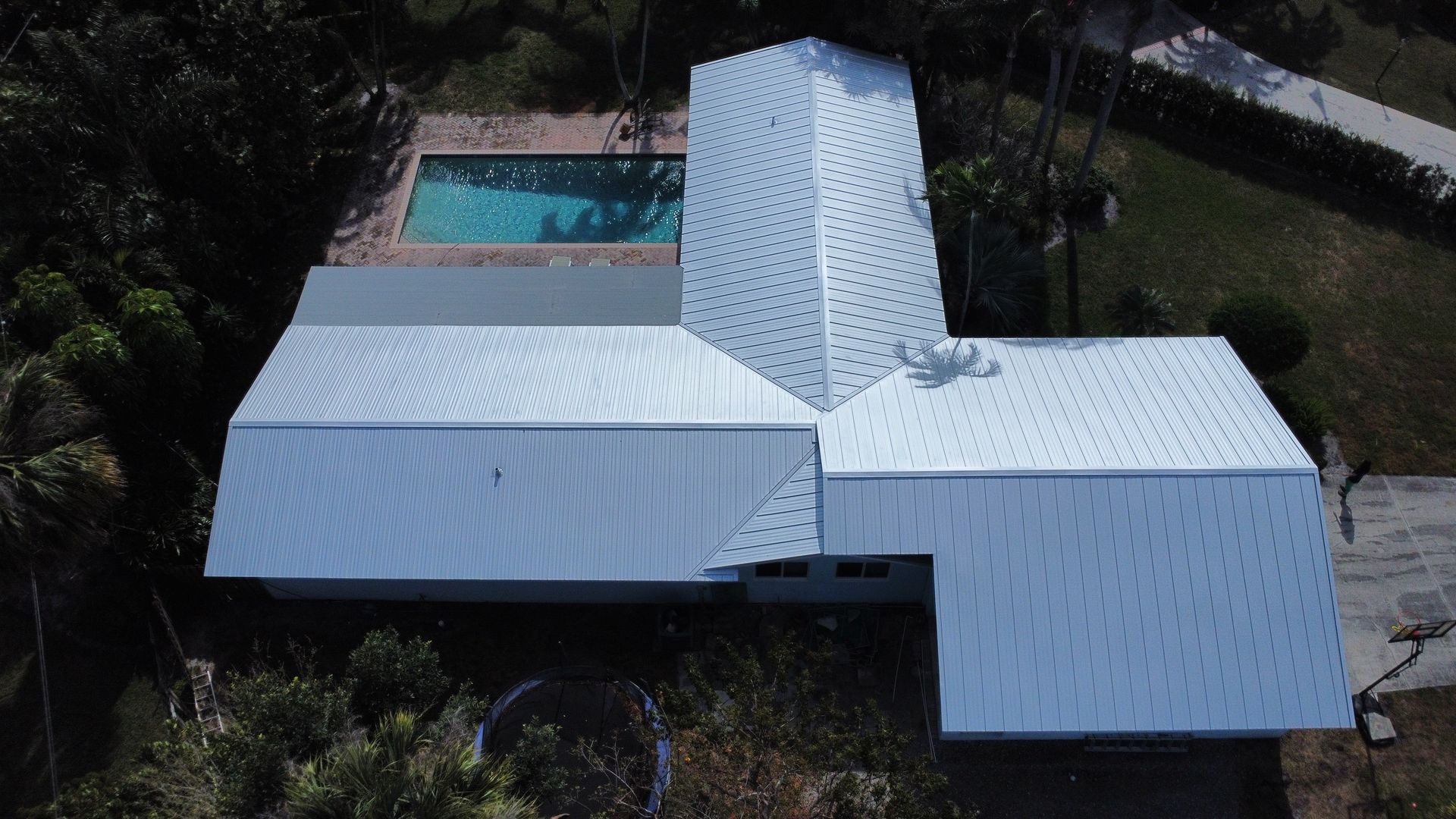 Aerial view of a house with a light metal roof beside a swimming pool and trees
