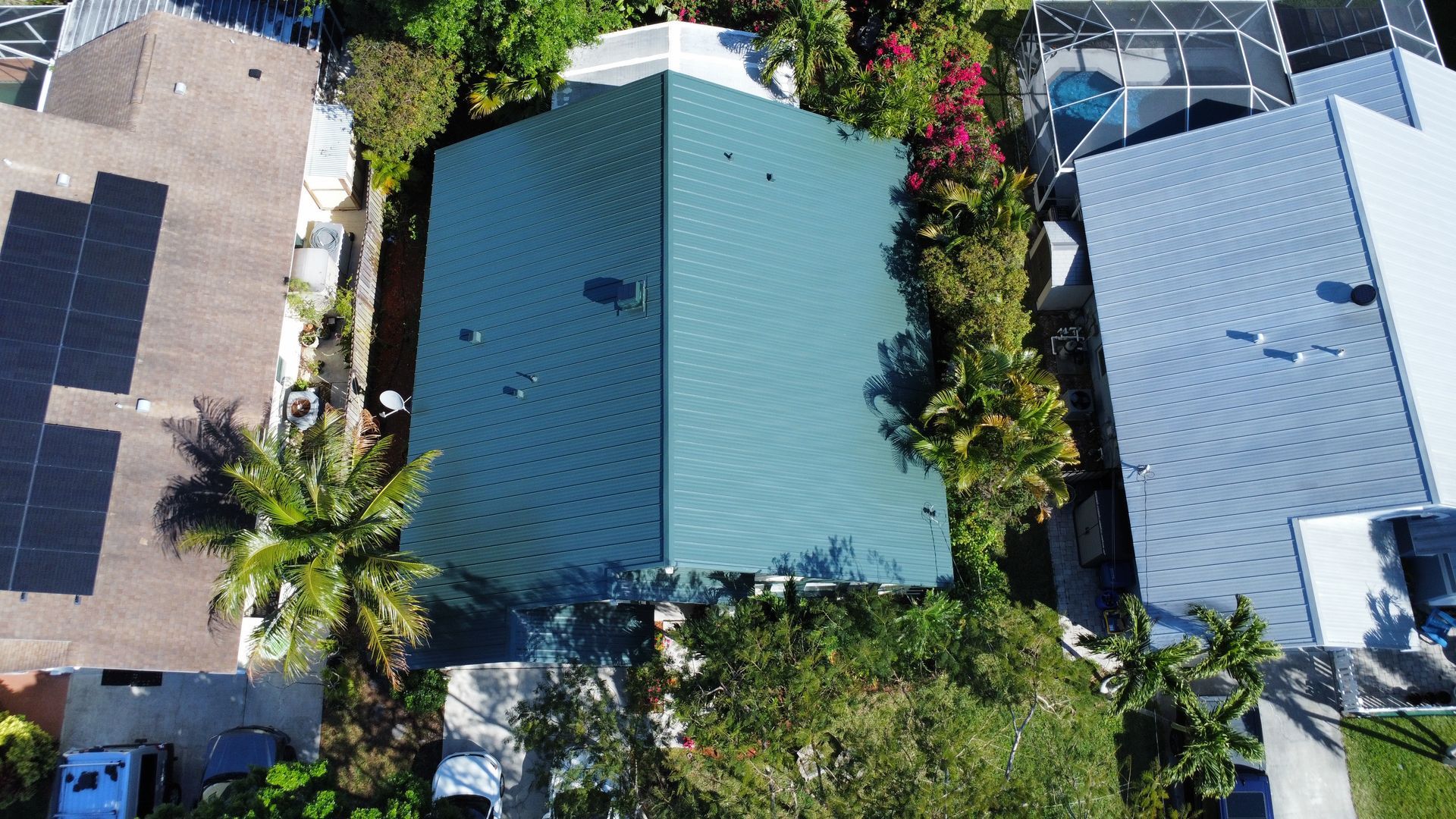 Aerial view of a teal-roofed house surrounded by palm trees and neighboring homes in a tropical neighborhood