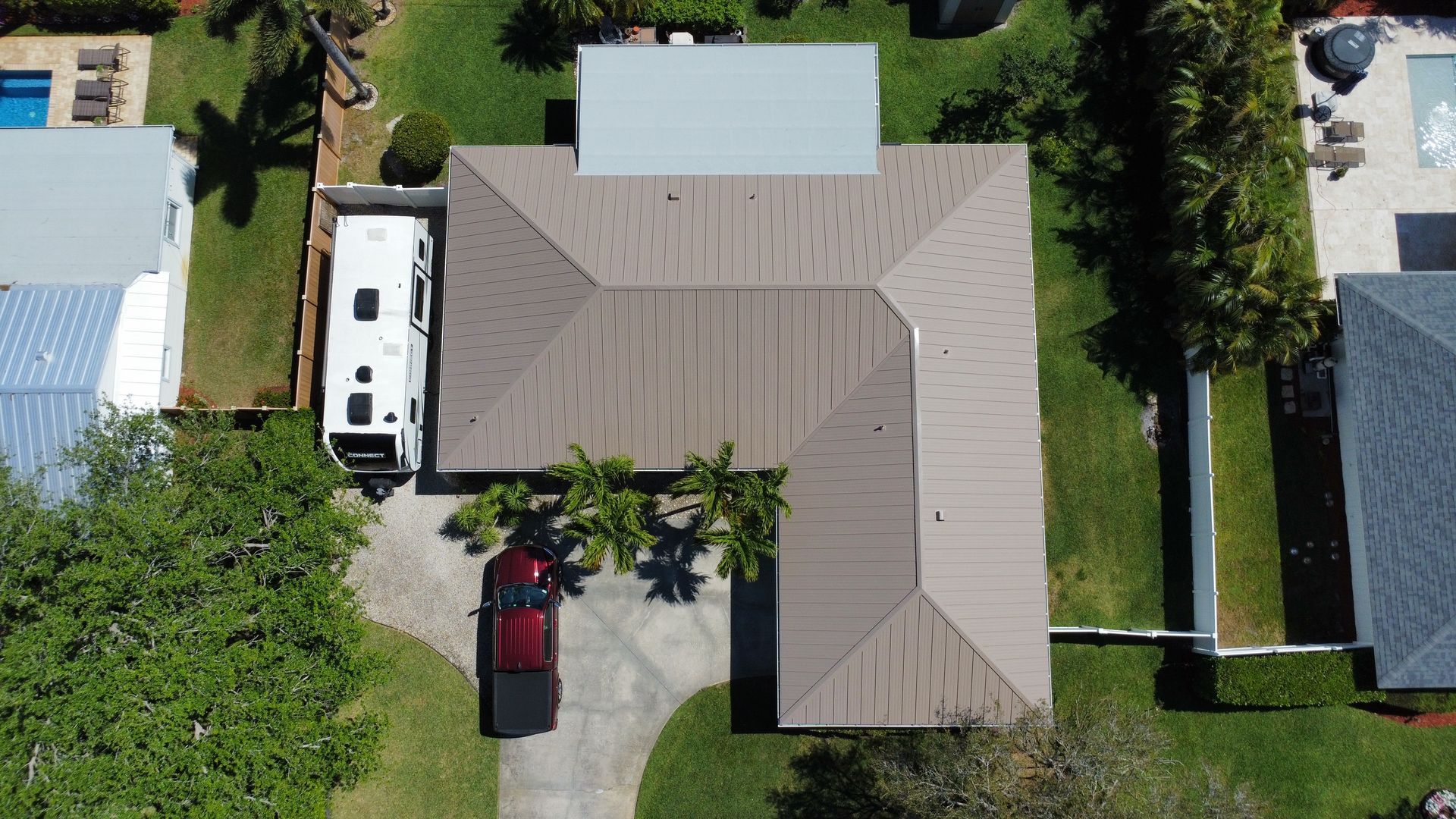 Aerial view of a tan-roofed house with a driveway, parked white van, red car, and surrounding trees.