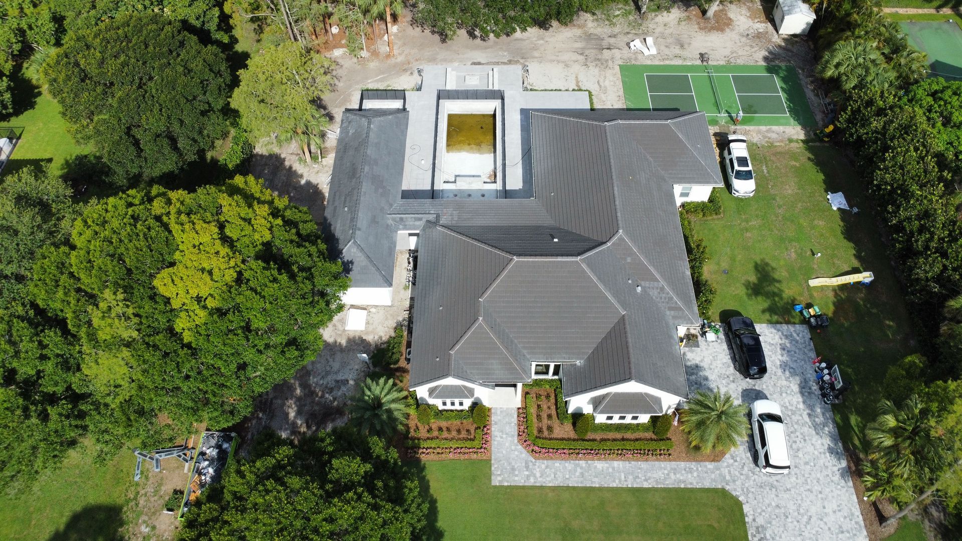Aerial view of a large house with a gray roof, surrounding lawn, driveway, and parked cars.