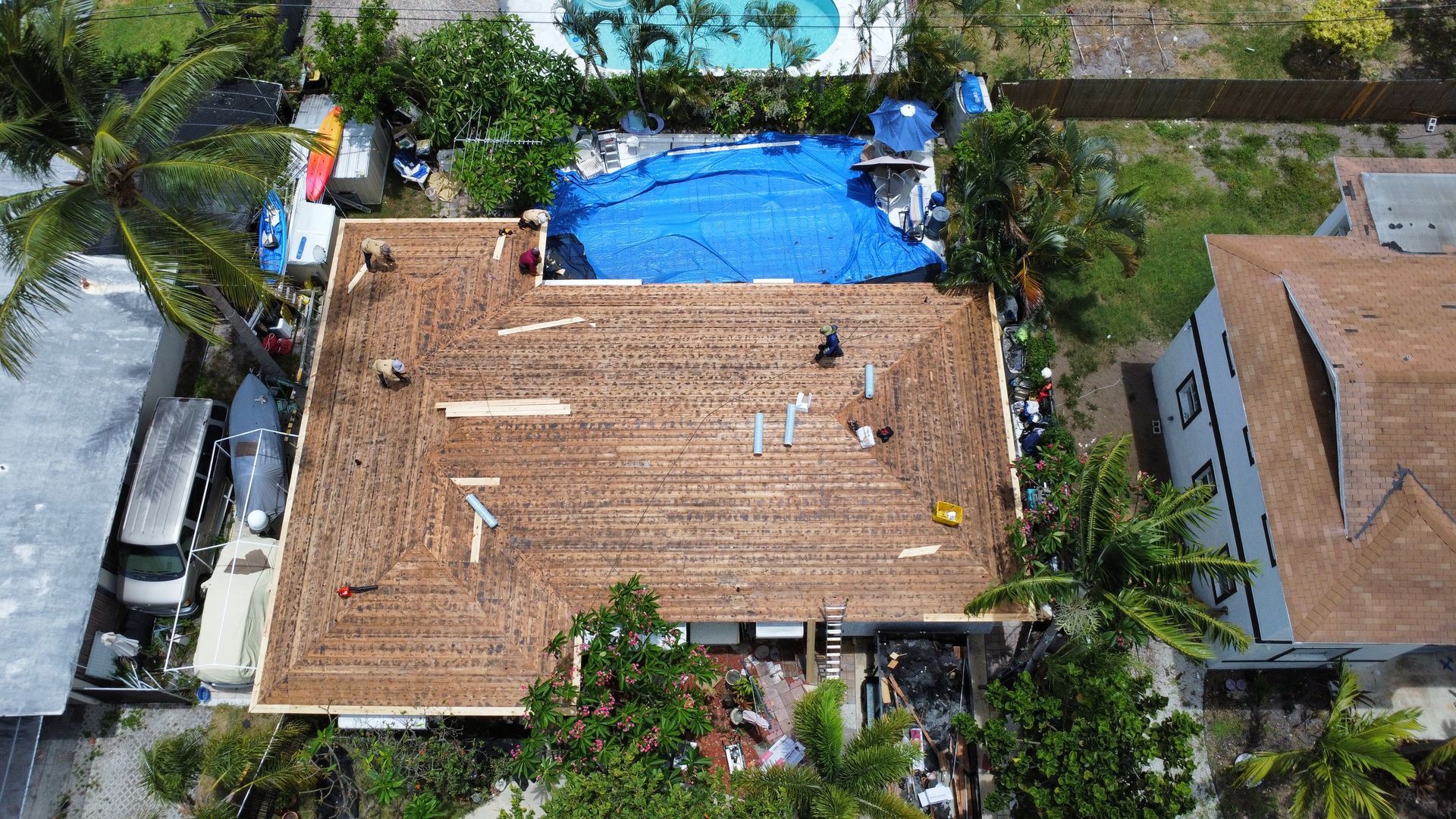 Aerial view of a house roof with workers repairing it beside a blue tarp-covered area.