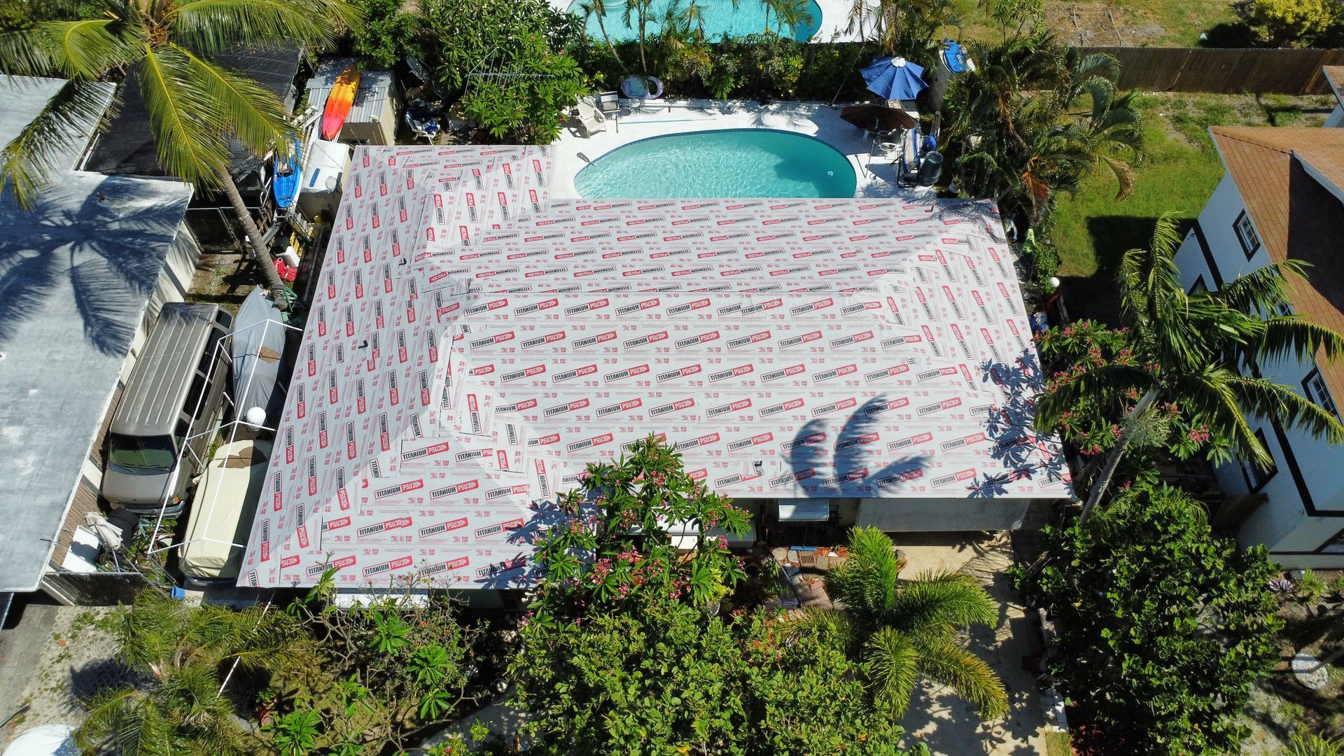 Aerial view of a house with a red-and-white roof beside a backyard pool and lush tropical trees.