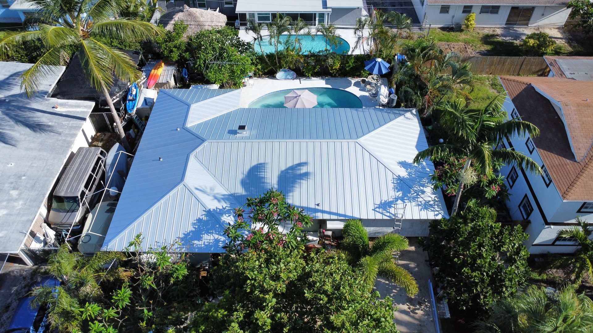 Aerial view of a blue-roofed house with a backyard pool, surrounded by trees and neighboring homes.