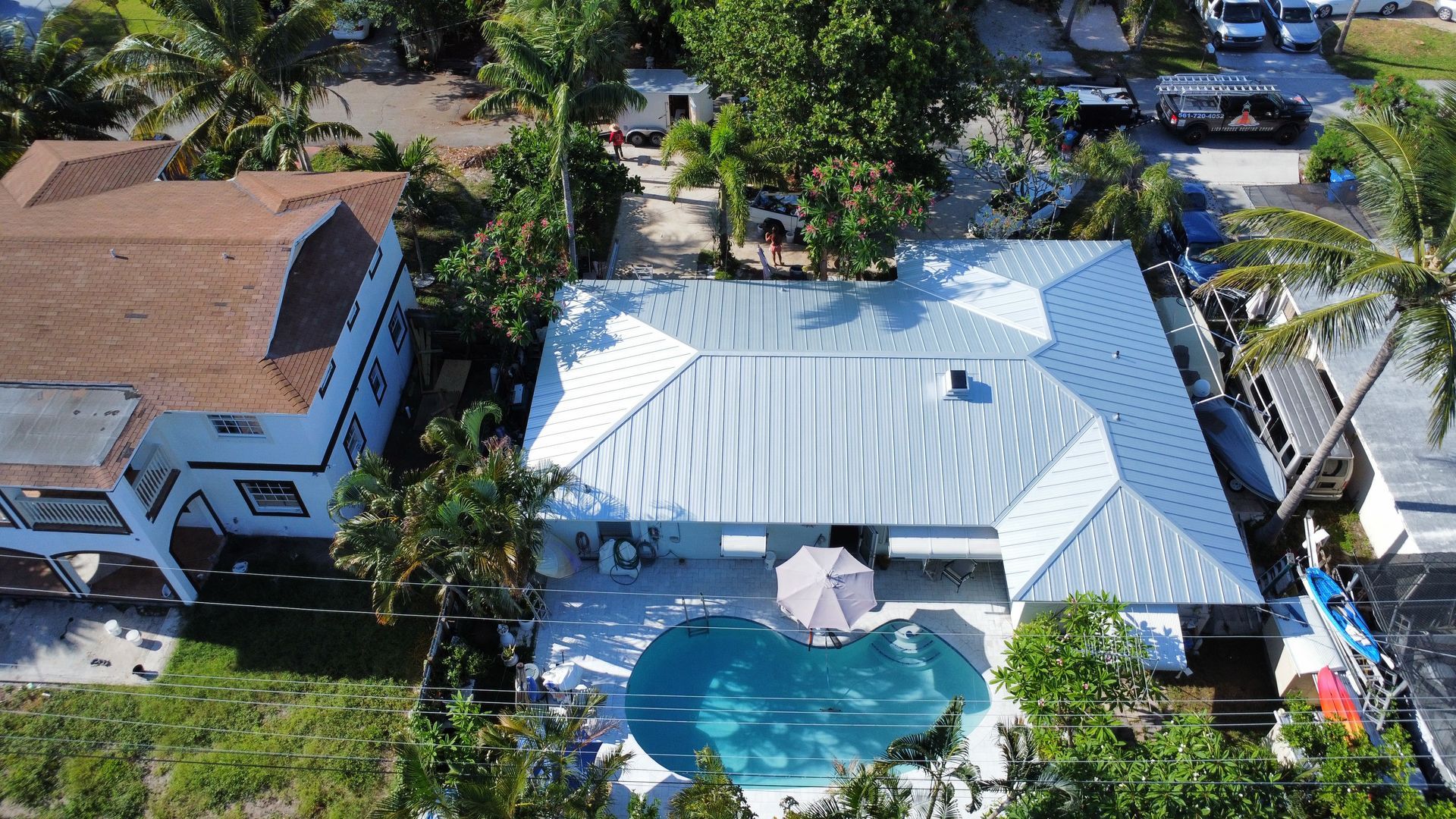 Aerial view of a white-roofed house with a backyard pool, surrounded by palm trees and neighboring homes