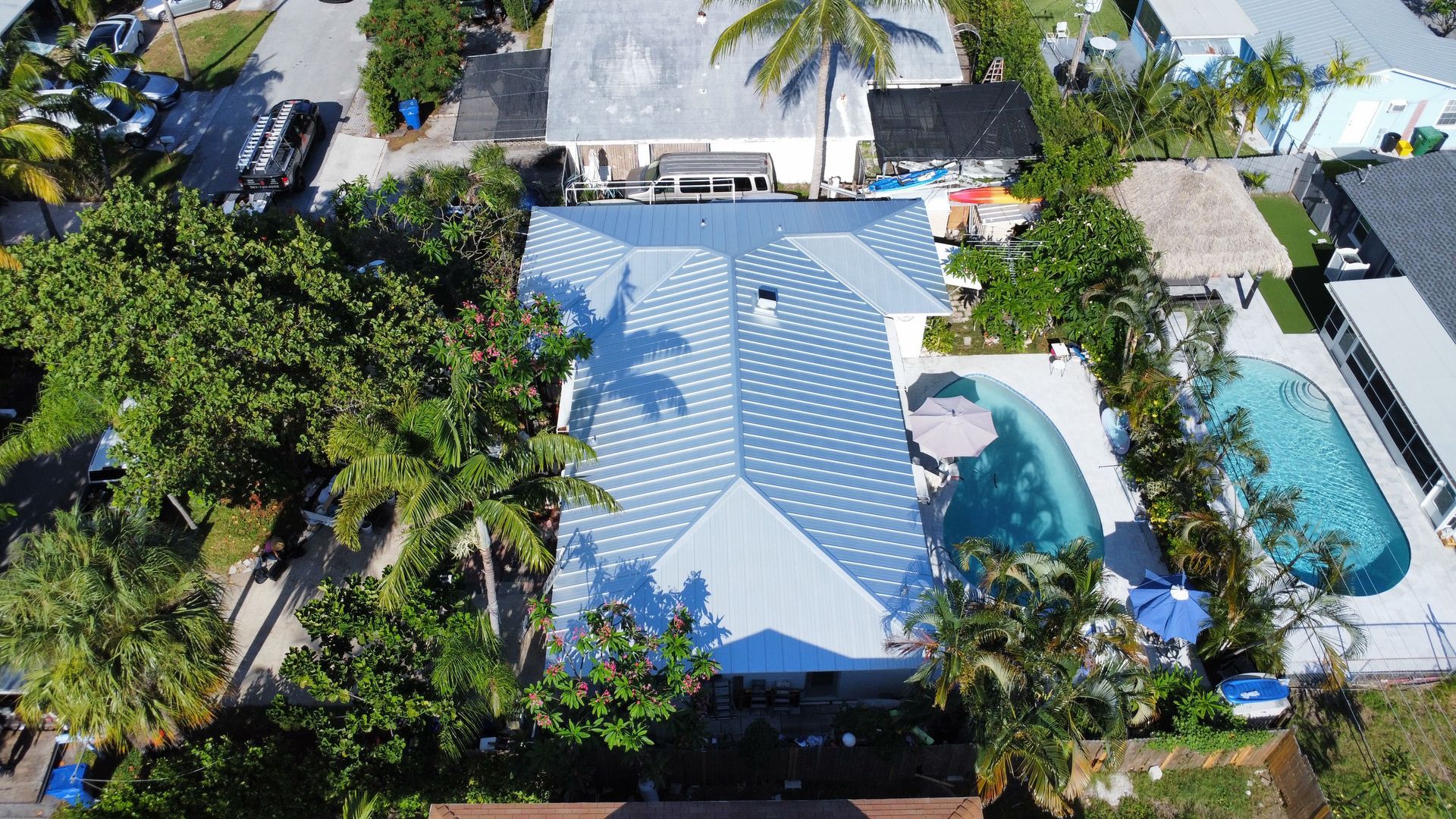 Aerial view of a blue-roofed house with a backyard pool, surrounded by palm trees and lush greenery