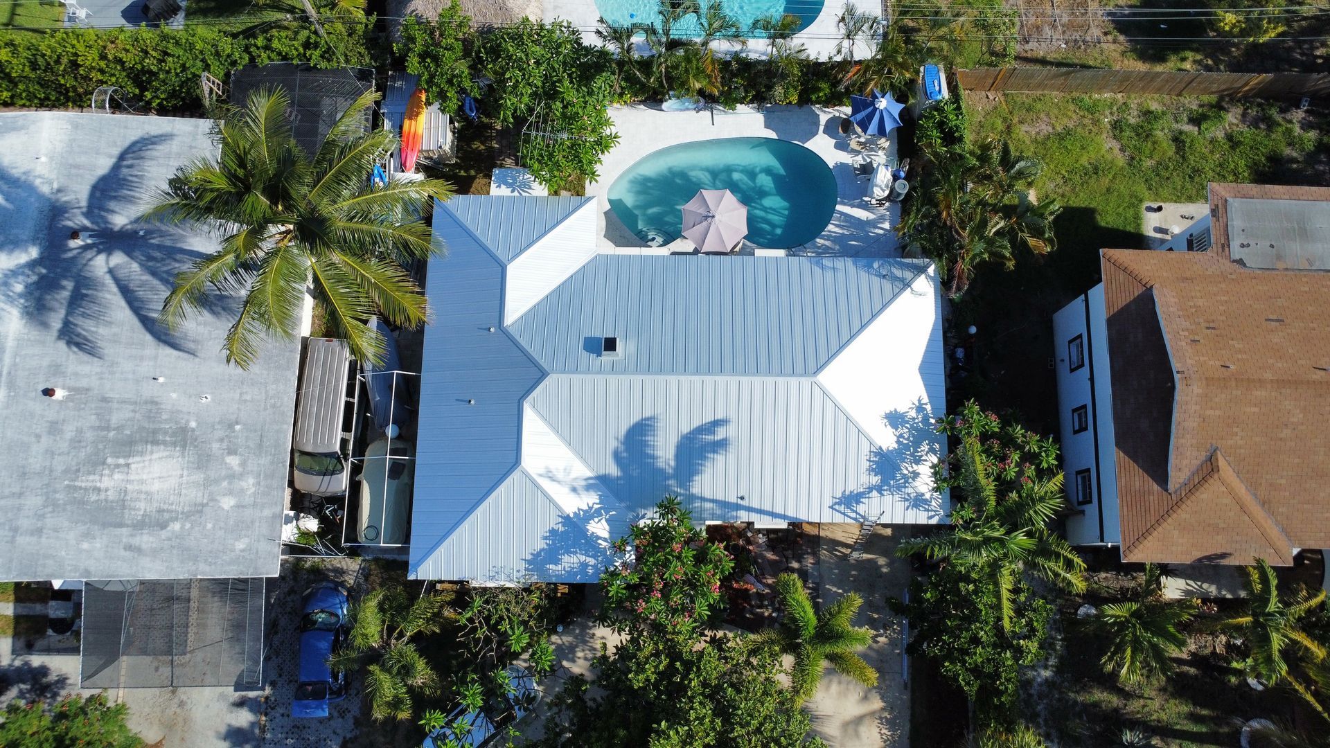 Aerial view of a house with a white roof, pool, and surrounding palm trees in a tropical neighborhood