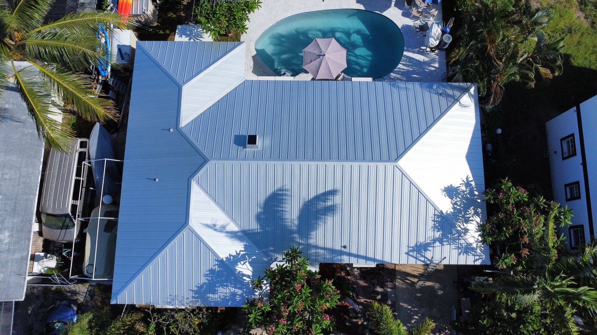 Aerial view of a blue-roofed house beside a swimming pool and palm trees.