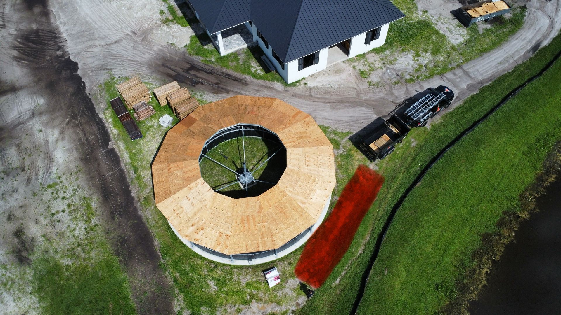 Aerial view of a circular wooden dock beside a house and grassy shoreline, with a parked vehicle nearby