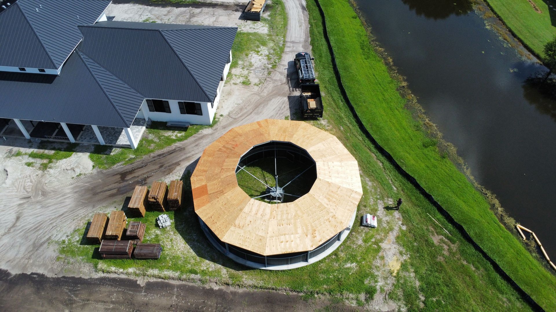 Aerial view of a round wooden pavilion beside a house, with picnic tables and a river nearby.