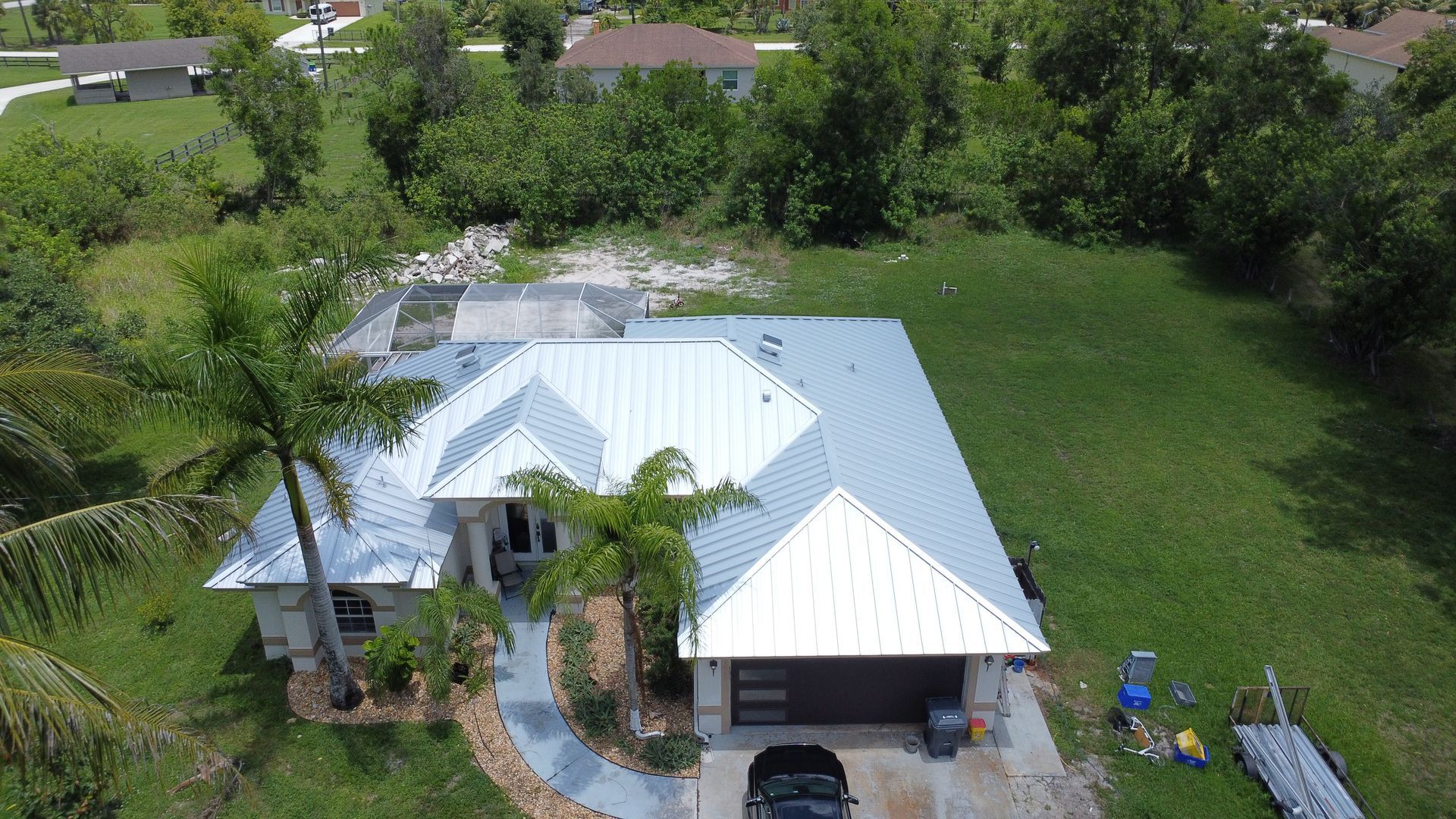 Aerial view of a white-roofed house with a driveway, palm trees, and a large green yard.